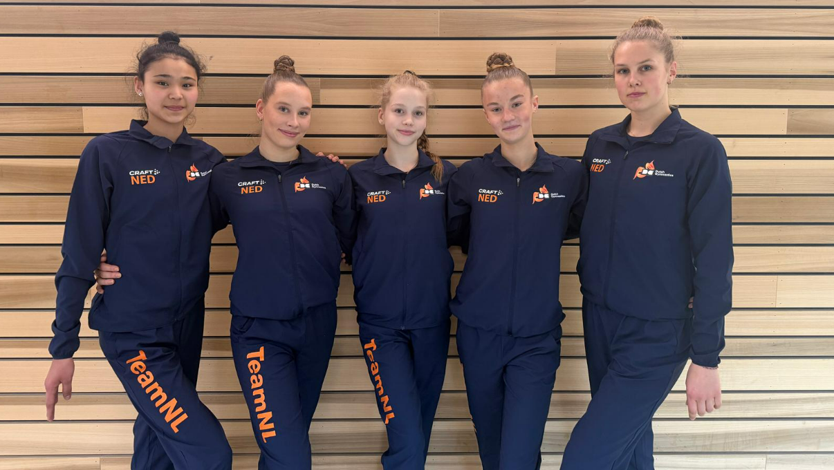 Vijf meisjes in blauwe sportkleding met "TeamNL" aan de zijkant, tegen een houten wand geposeerd.