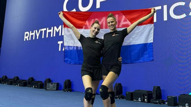 Twee vrouwen poseren glimlachend met een Nederlandse vlag op een podium met grote tekst op de achtergrond.