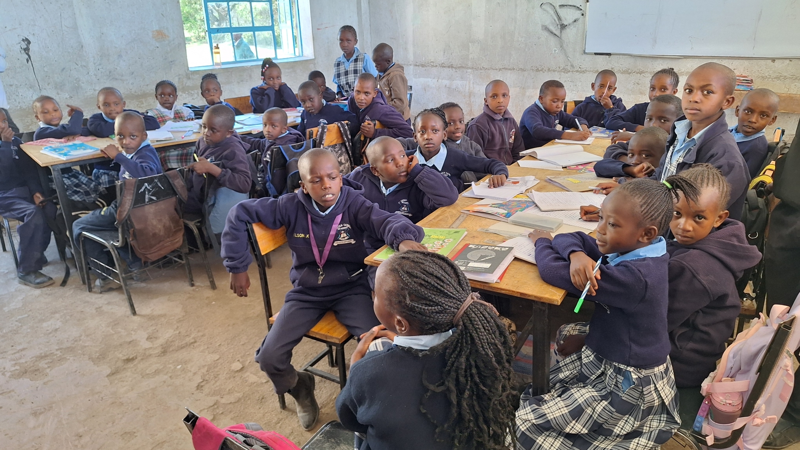 Kinderen in schooluniform zitten in een klaslokaal met boeken en schriften op tafels, luisterend naar instructies.