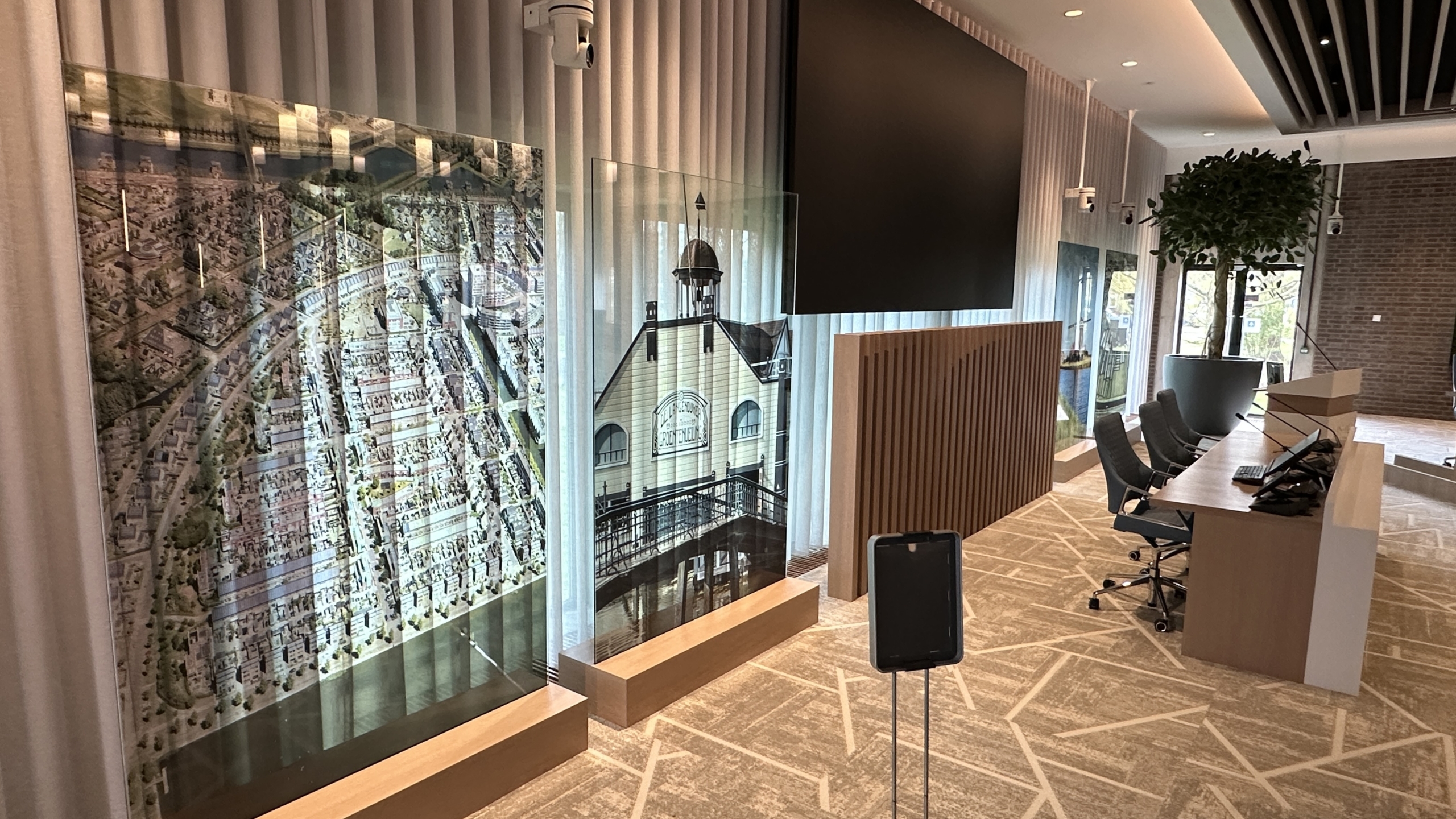 Moderne vergaderruimte met stoelen, tafels, een grote monitor, glazen afbeeldingen aan de muur en een grote kamerplant.