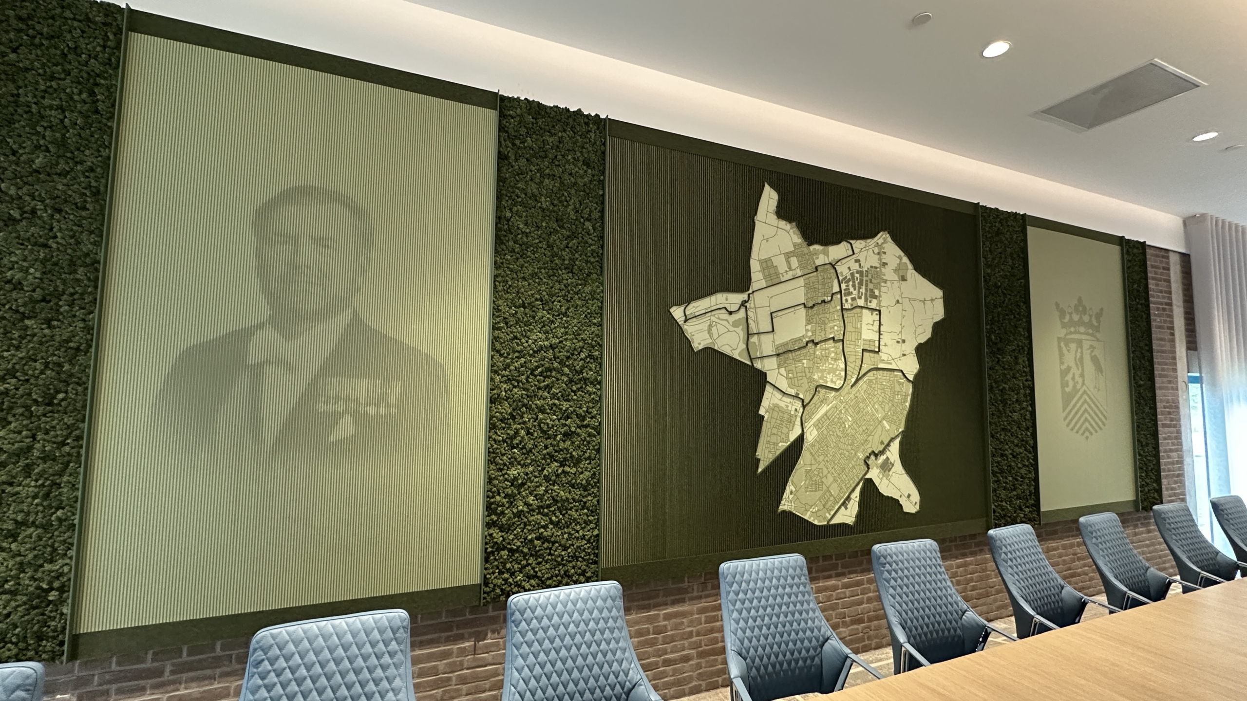Vergaderruimte met een grote kaart, portret en wapenschild op de muur, en blauwe stoelen rond een houten tafel.