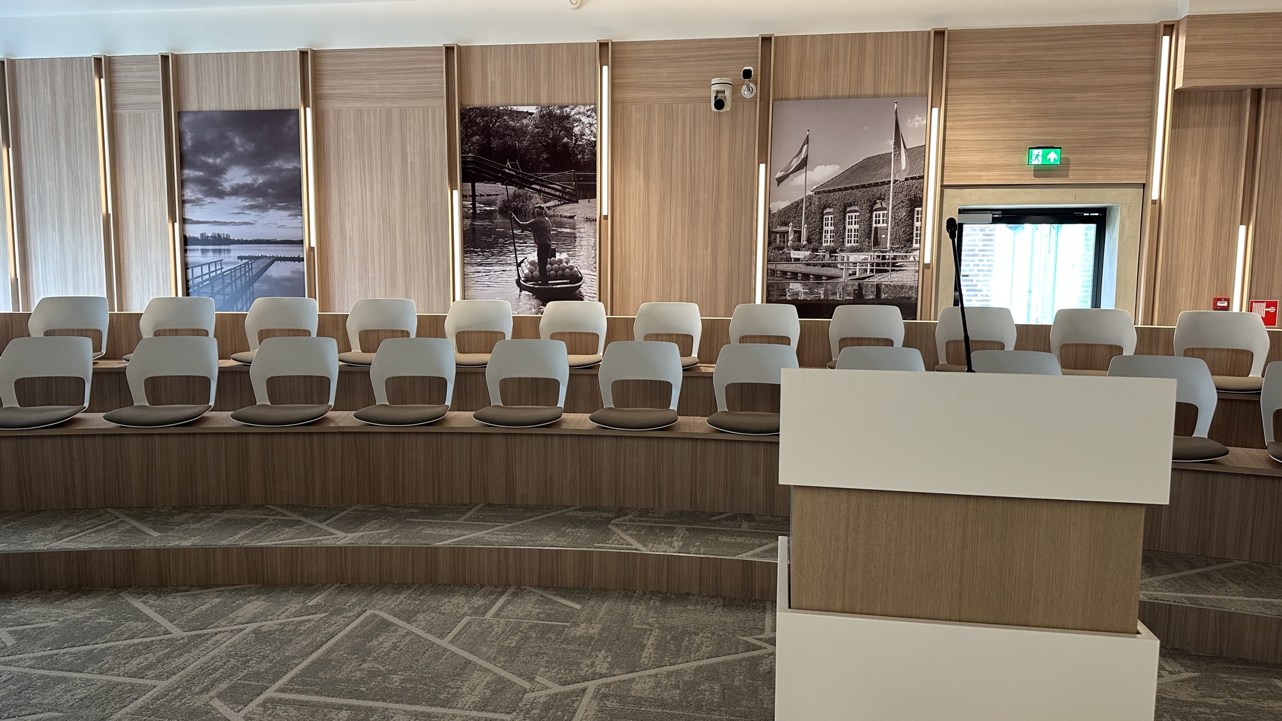 Lege conferentiezaal met houten afwerking, meerdere rijen moderne stoelen, en historische zwart-wit foto's aan de muur.