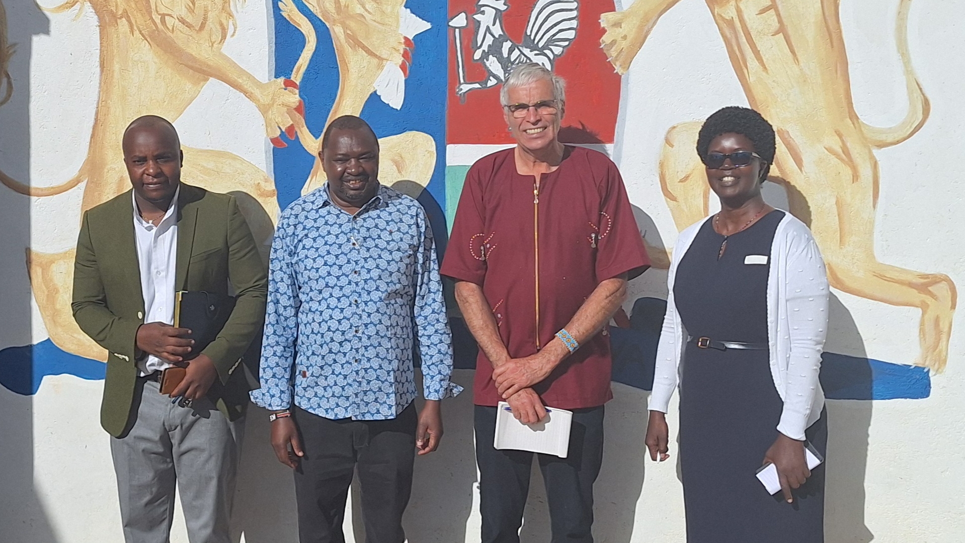 Vier mensen poseren voor een muur met een schildering van leeuwen en een wapenschild. Ze glimlachen bij zonnig weer.