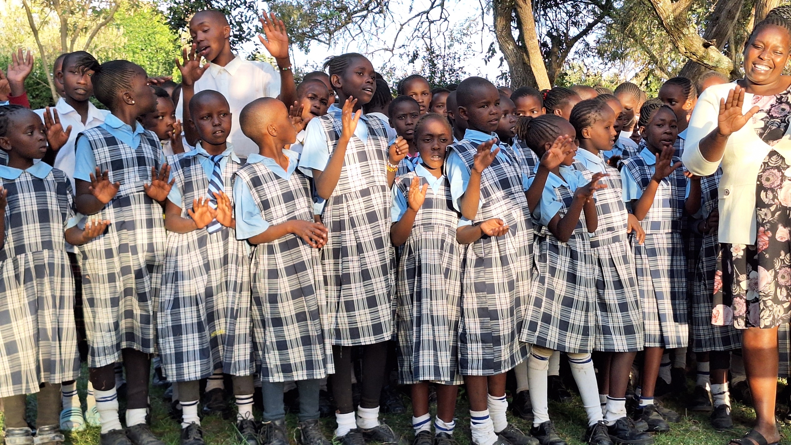 Een groep schoolkinderen in schooluniform zingt in een buitenomgeving, begeleid door een man en een vrouw.