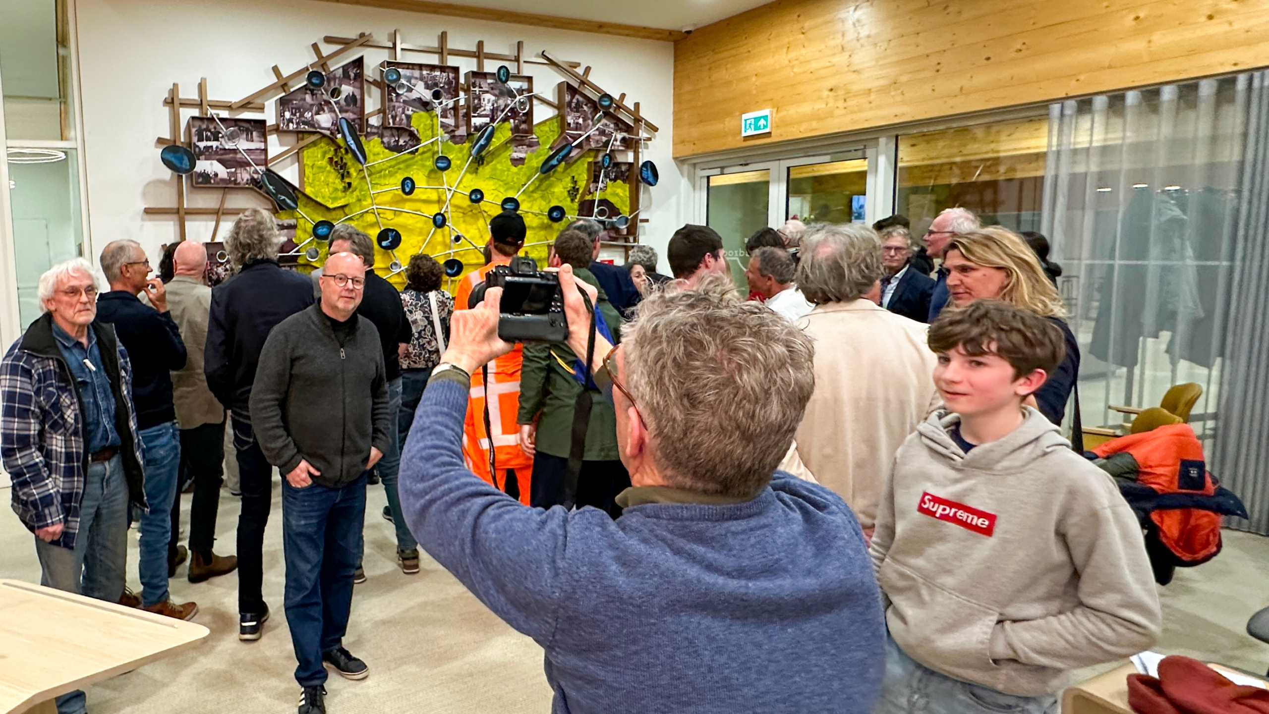 Mensen verzamelen zich rond een wandkunstwerk in een moderne ruimte, terwijl iemand een foto maakt.