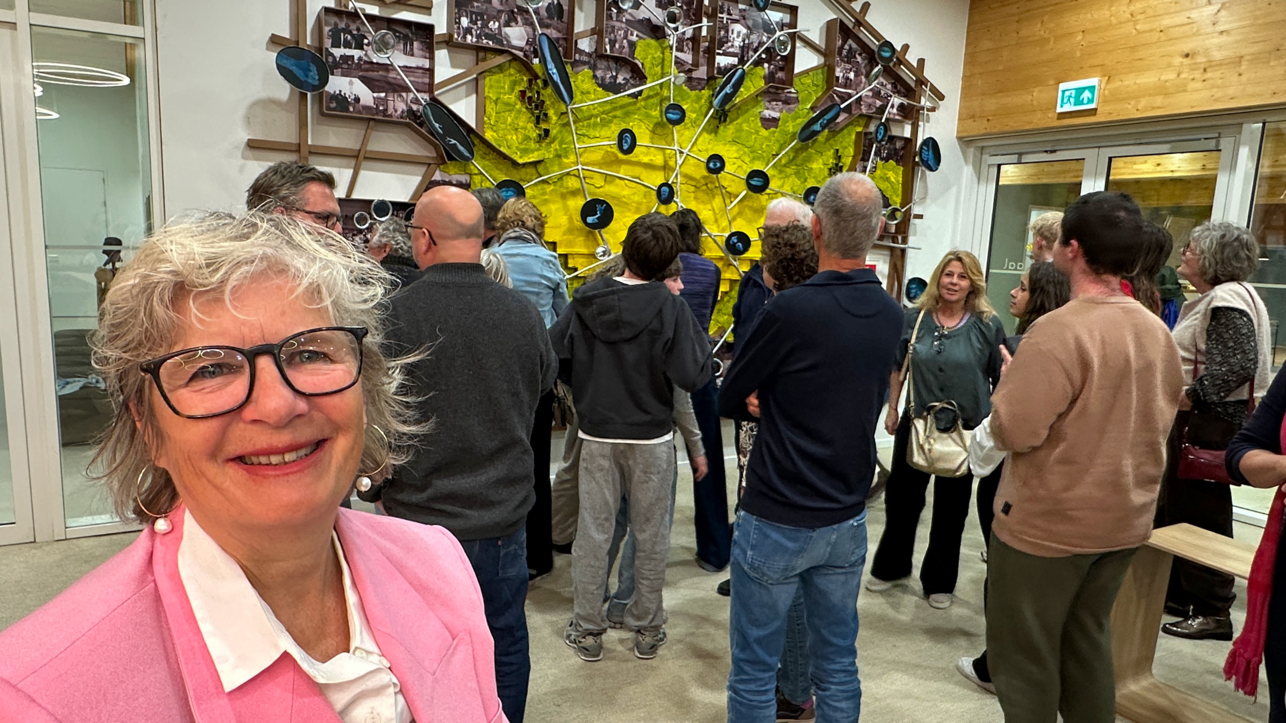 Mensen staan in een galerij, met kleurrijke kunstwerken aan de muur; een vrouw in een roze jas lacht naar de camera.