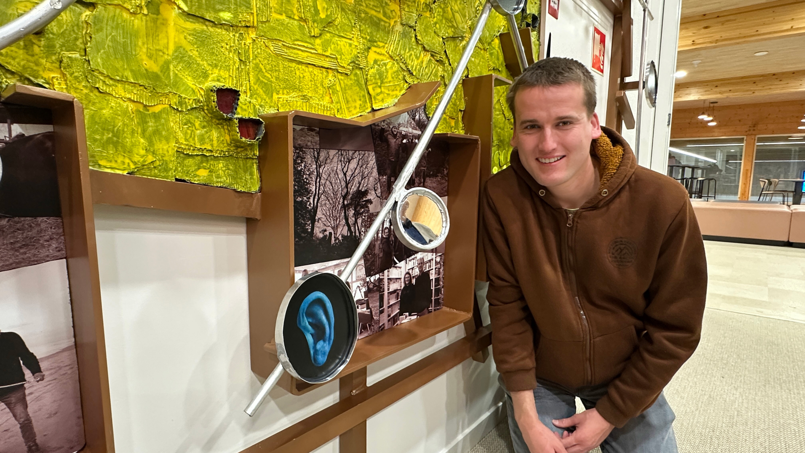 Een man knielt naast een kunstwerk met een gele achtergrond en foto's, inclusief een blauwe afbeelding van een oor.