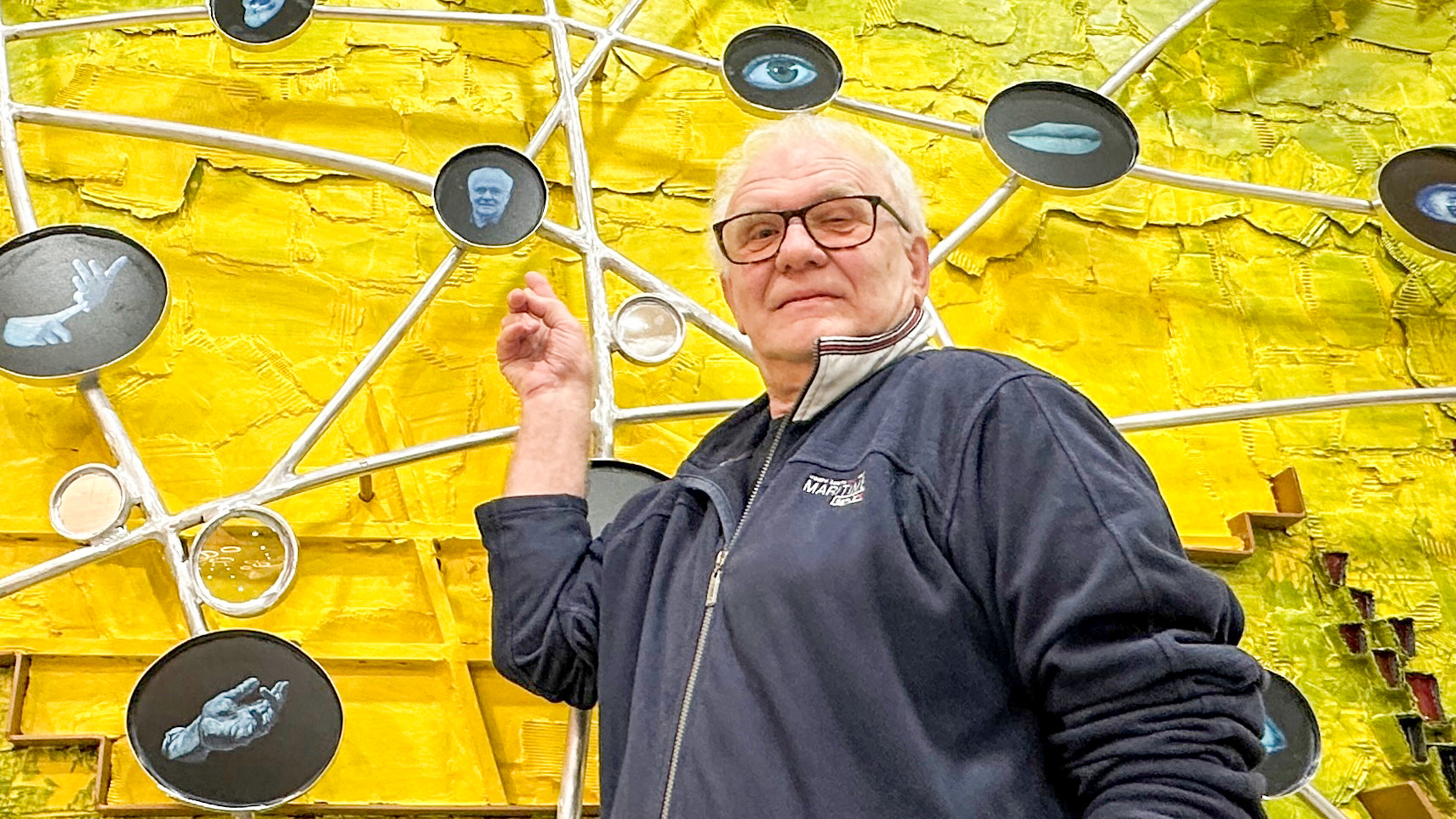 Man met bril in donker jack voor een kunstwerk met gele achtergrond en verbonden fotocirkels.