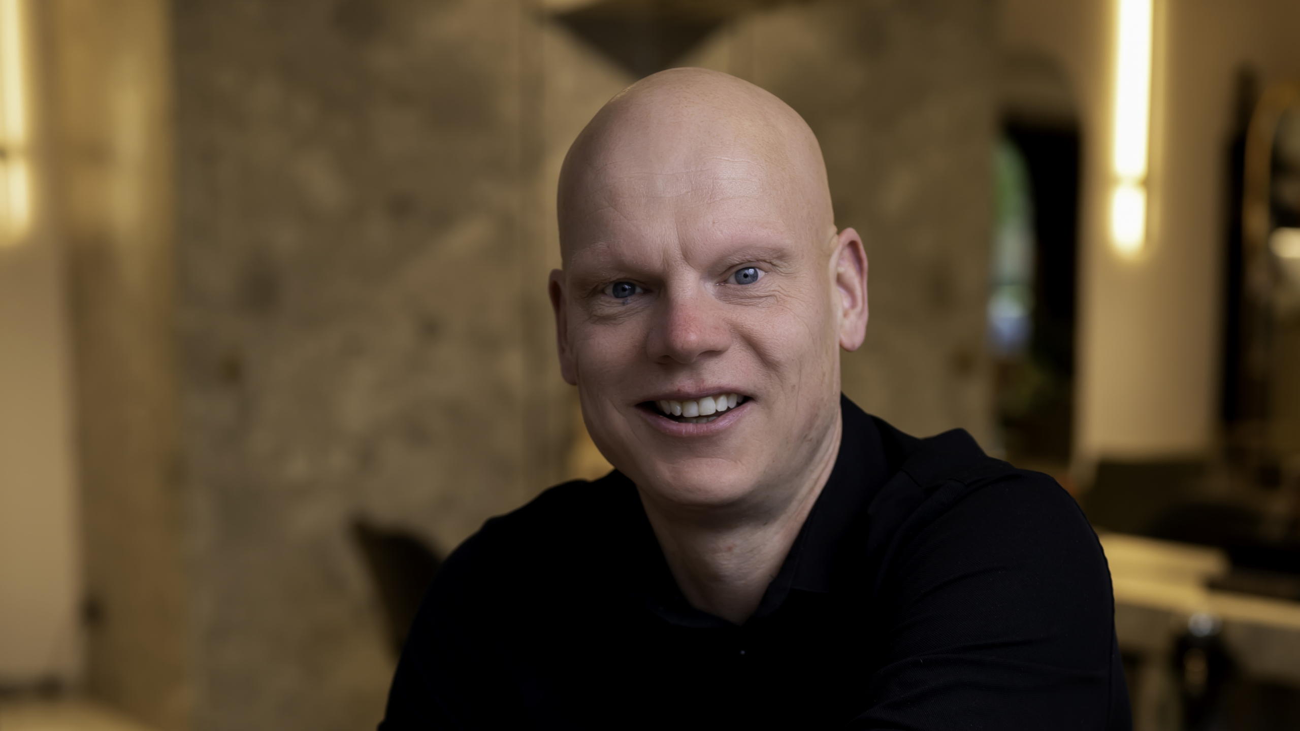 Glimlachende man met een kaal hoofd en een zwarte trui in een warme, rustige omgeving.