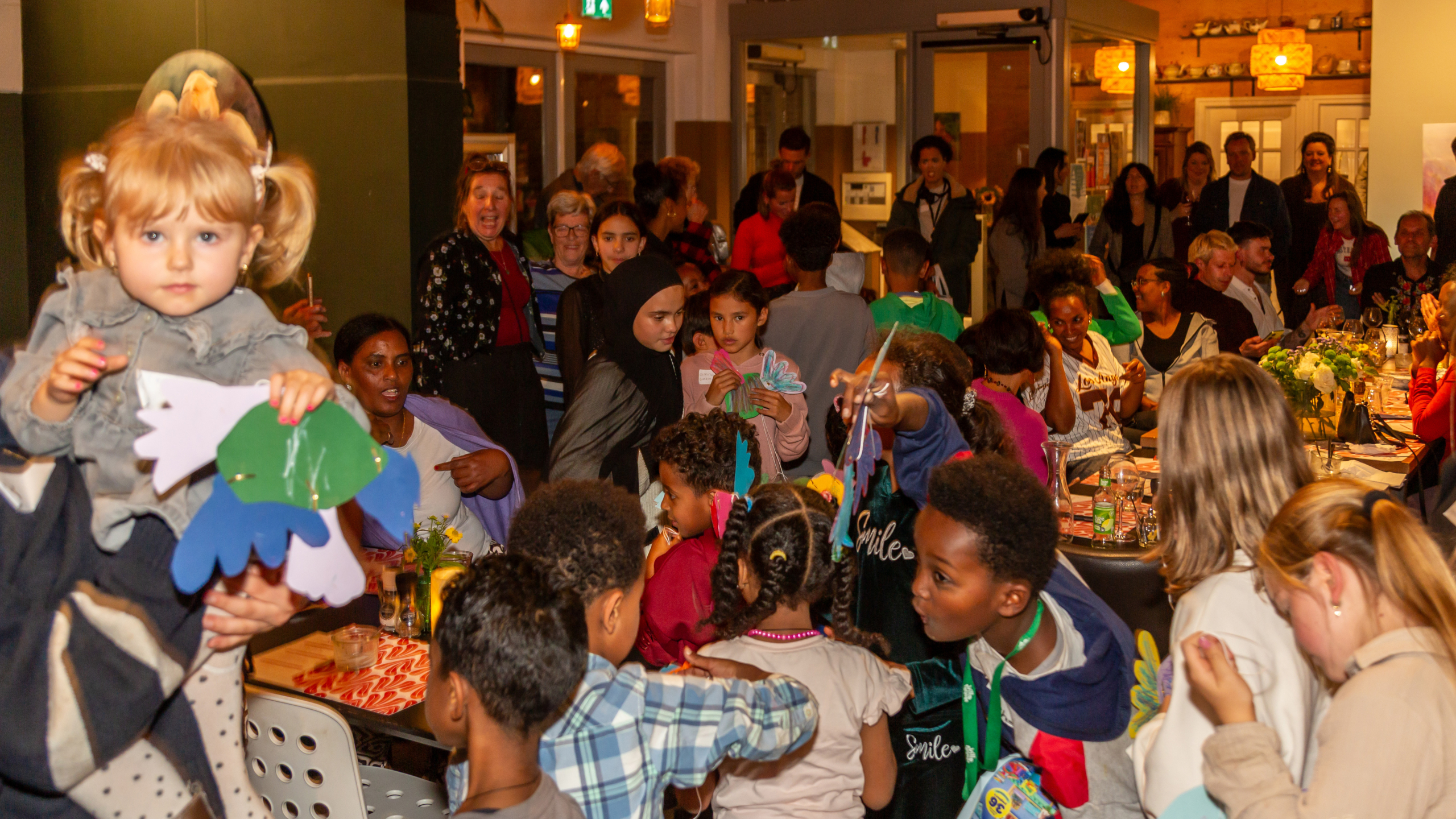 Groep mensen, waaronder kinderen, genieten van een drukbezochte bijeenkomst; een kind vooraan houdt gekleurd papier vast.