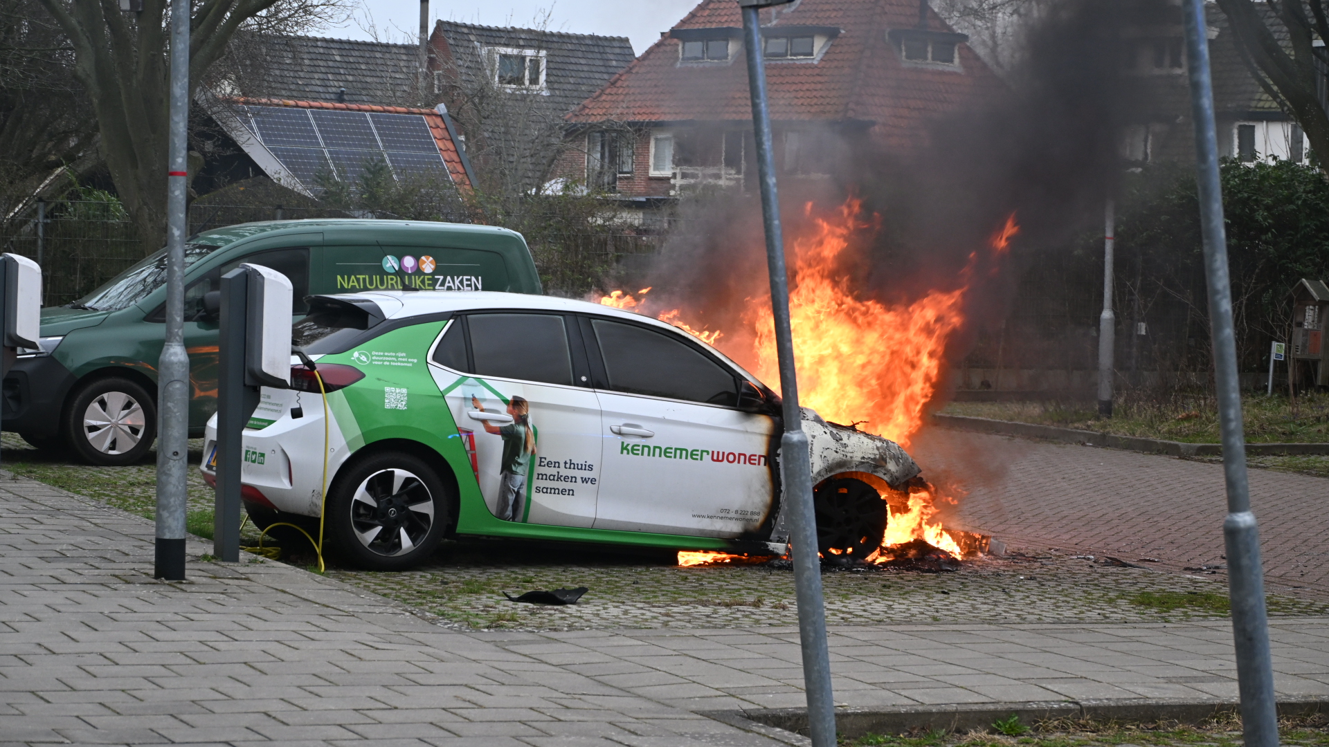 Een geparkeerde auto staat in brand met vlammen uit de motorkap, terwijl een ander voertuig en gebouwen op de achtergrond zichtbaar zijn.
