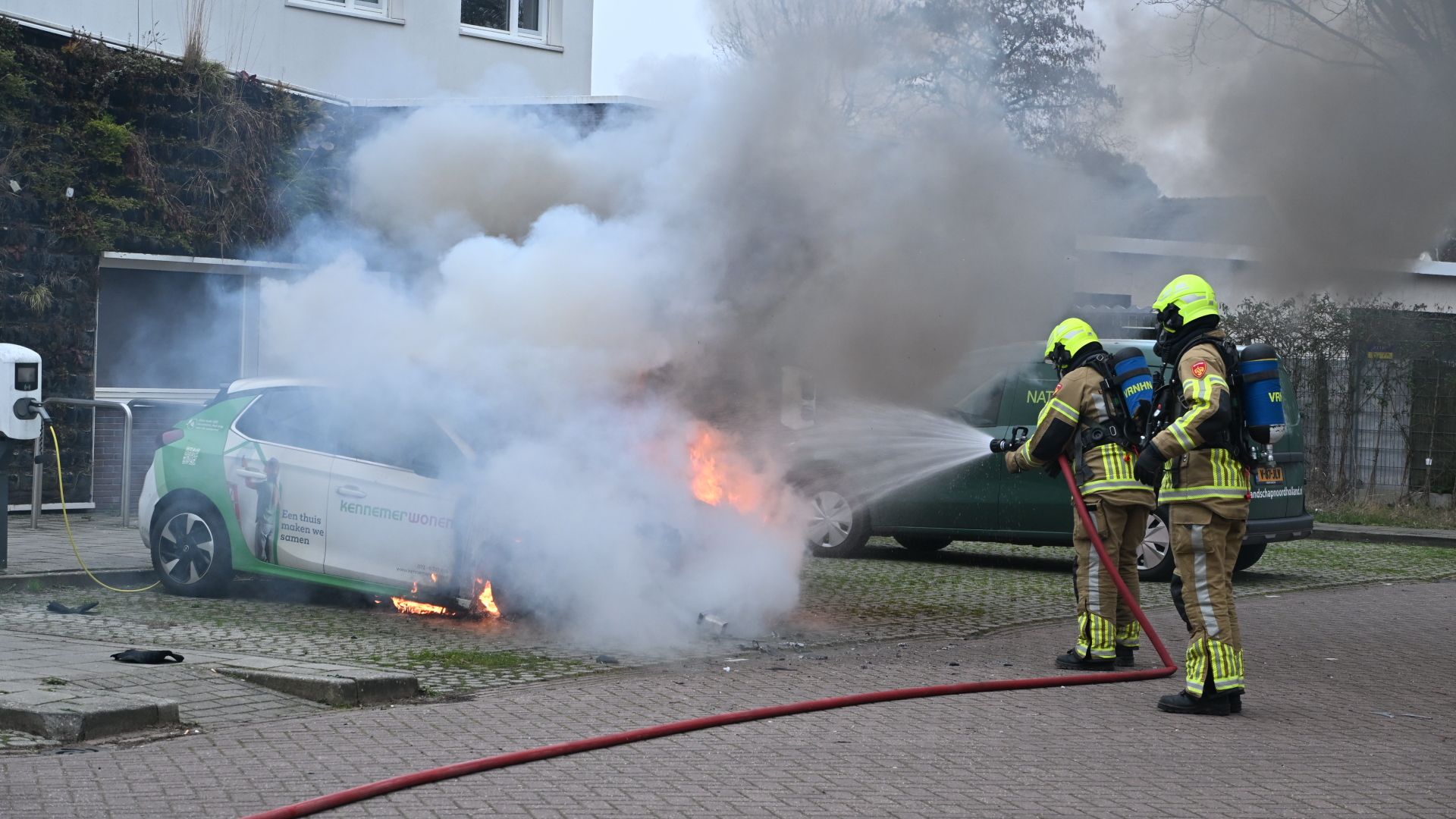 Brandweerlieden blussen een elektrische auto die in brand staat bij een laadpaal, omgeven door rook.