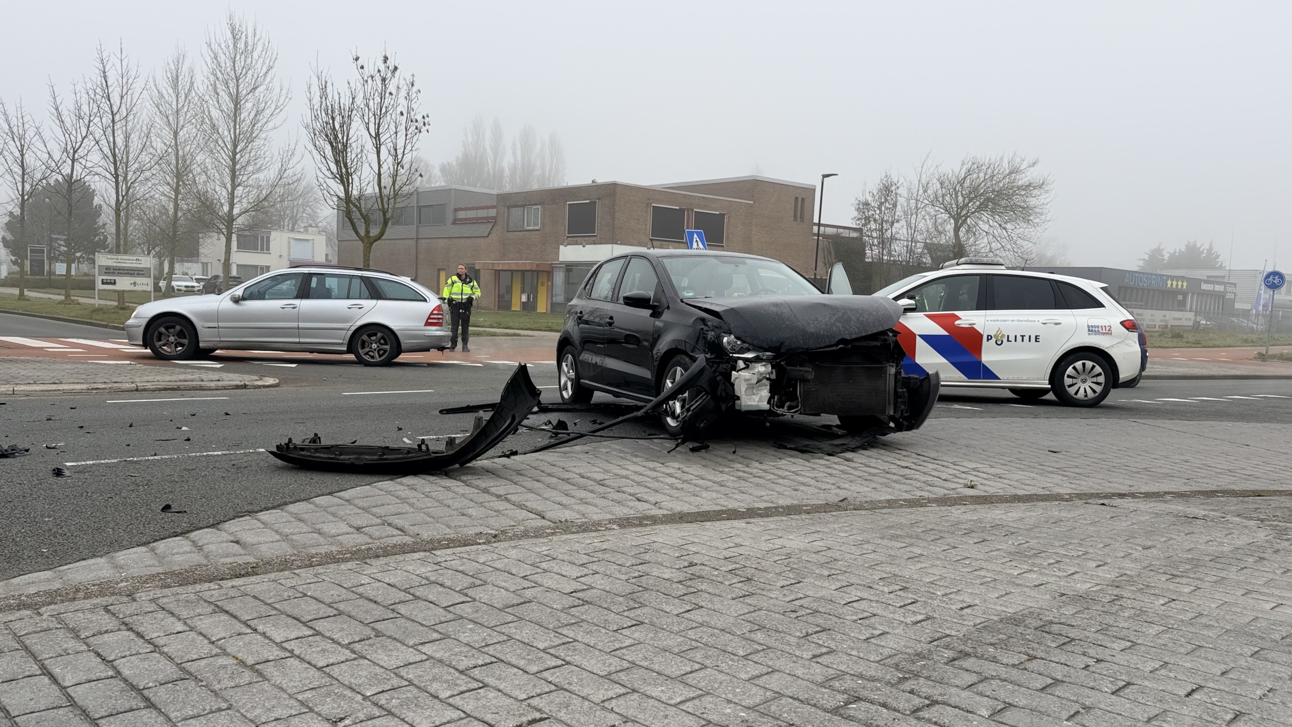 Auto met zware schade aan de voorzijde op een kruispunt, met politie en passerende voertuigen in de achtergrond.