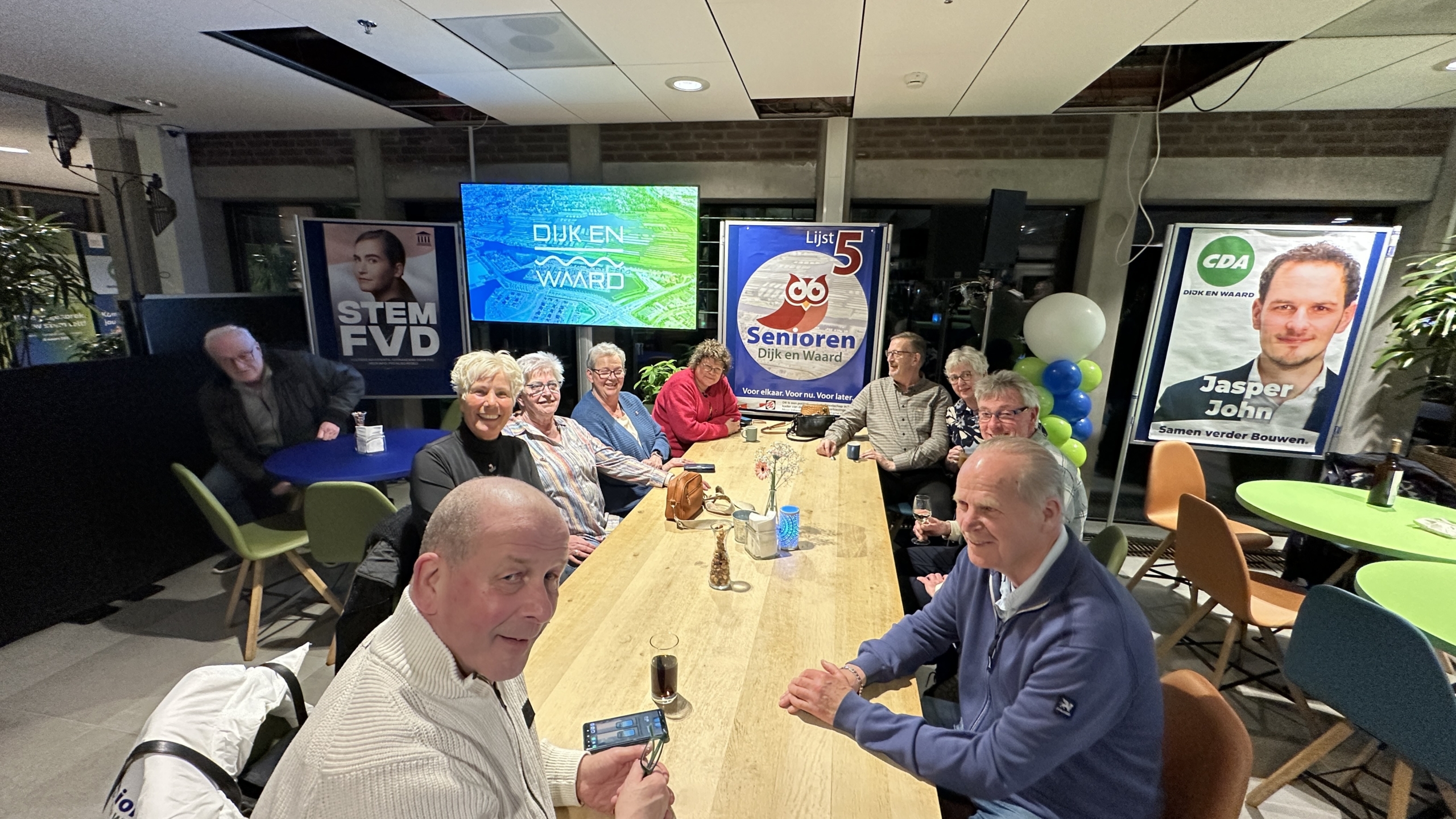 Groep mensen zit aan een tafel in een bijeenkomst, omringd door politieke posters en schermen.