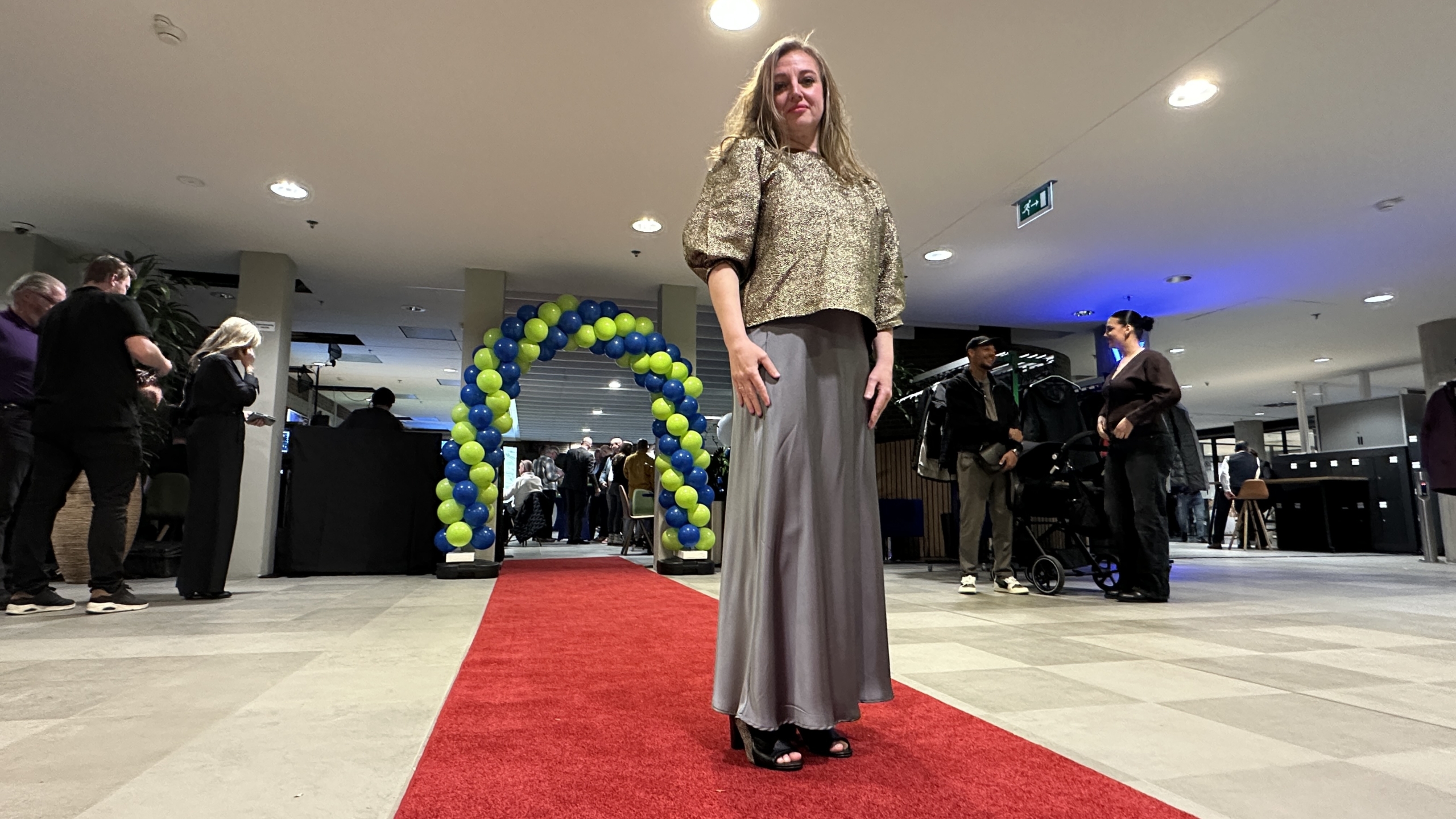 Vrouw in stijlvolle outfit staat op een rode loper met op de achtergrond een boog van blauwe en gele ballonnen.