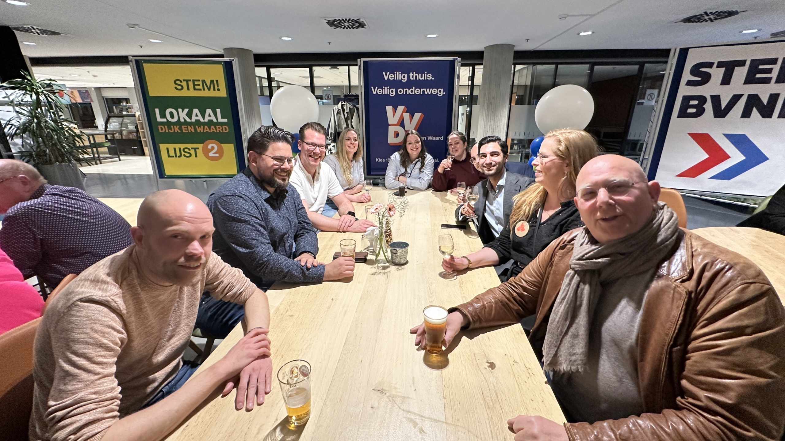 Mensen zitten rond een tafel met drankjes, omgeven door verkiezingsposters van verschillende politieke partijen.