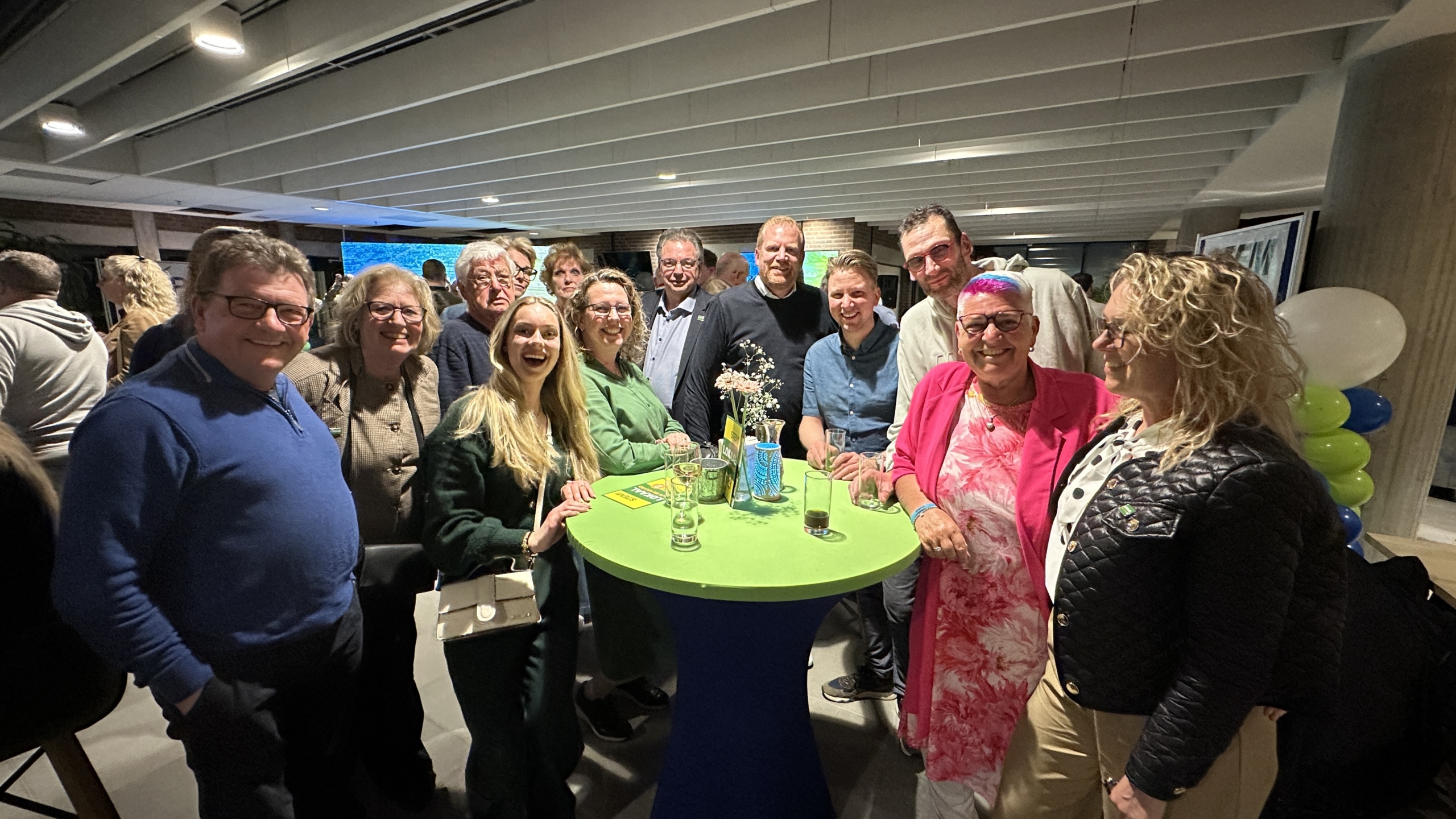 Groep mensen verzameld rond een tafel op een feestelijke bijeenkomst met kleurrijke ballonnen op de achtergrond.