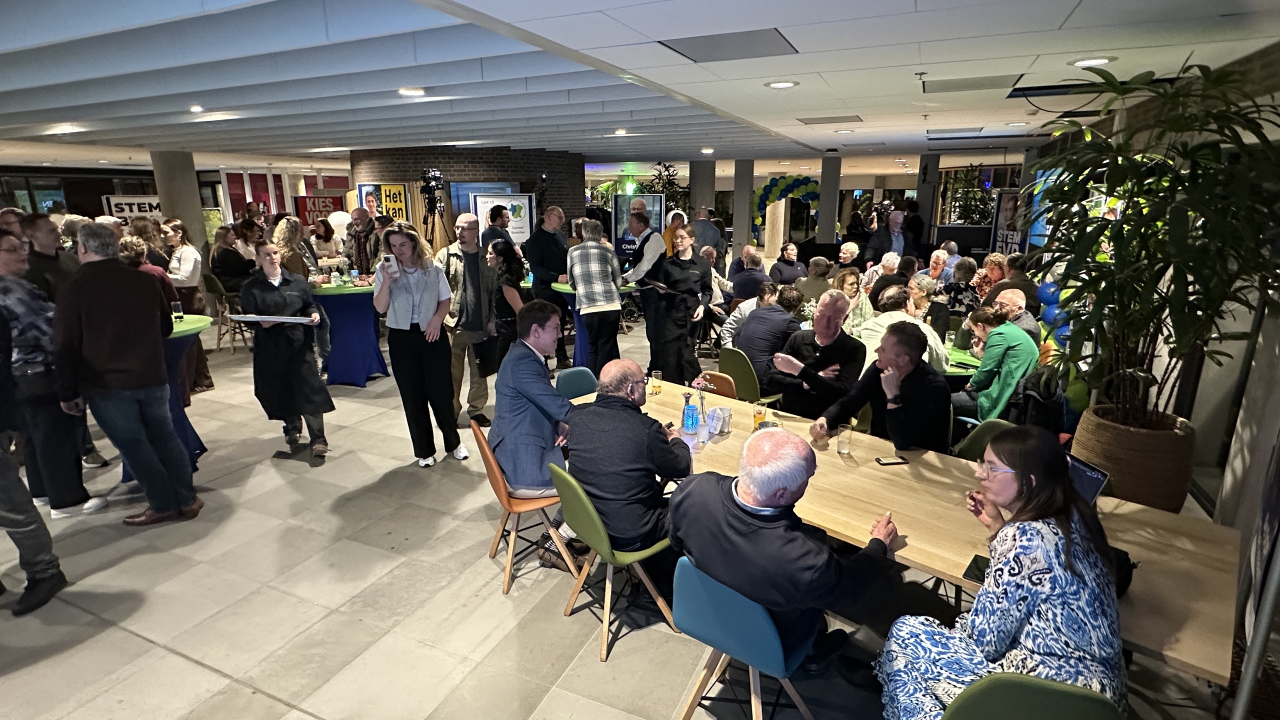 Een groep mensen op een bijeenkomst in een ruime, verlichte zaal met tafels en kleurrijke stoelen.