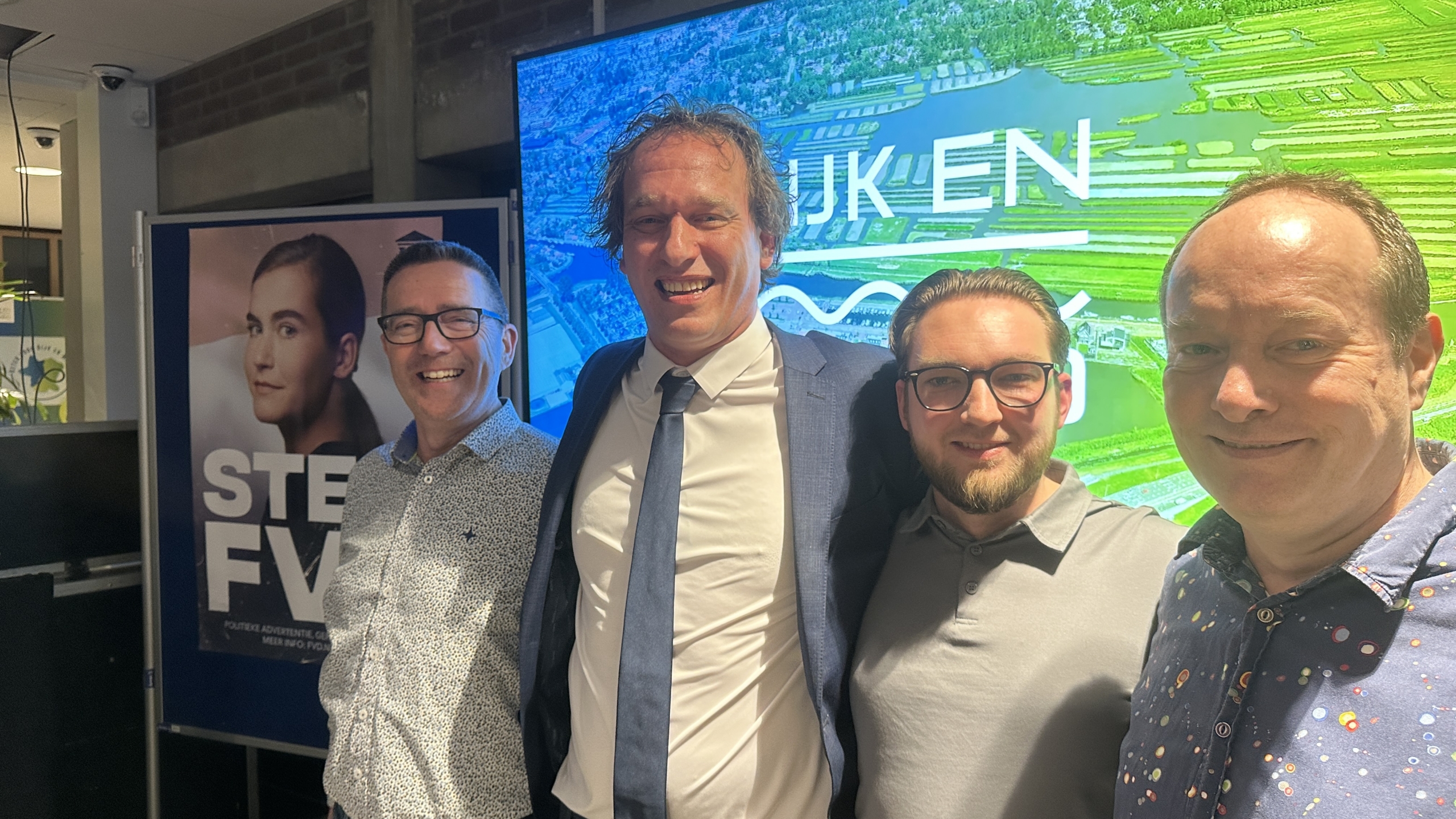 Vier mannen poseren samen voor een kleurrijk scherm en een poster met een gezicht en de tekst "STEM FVD".