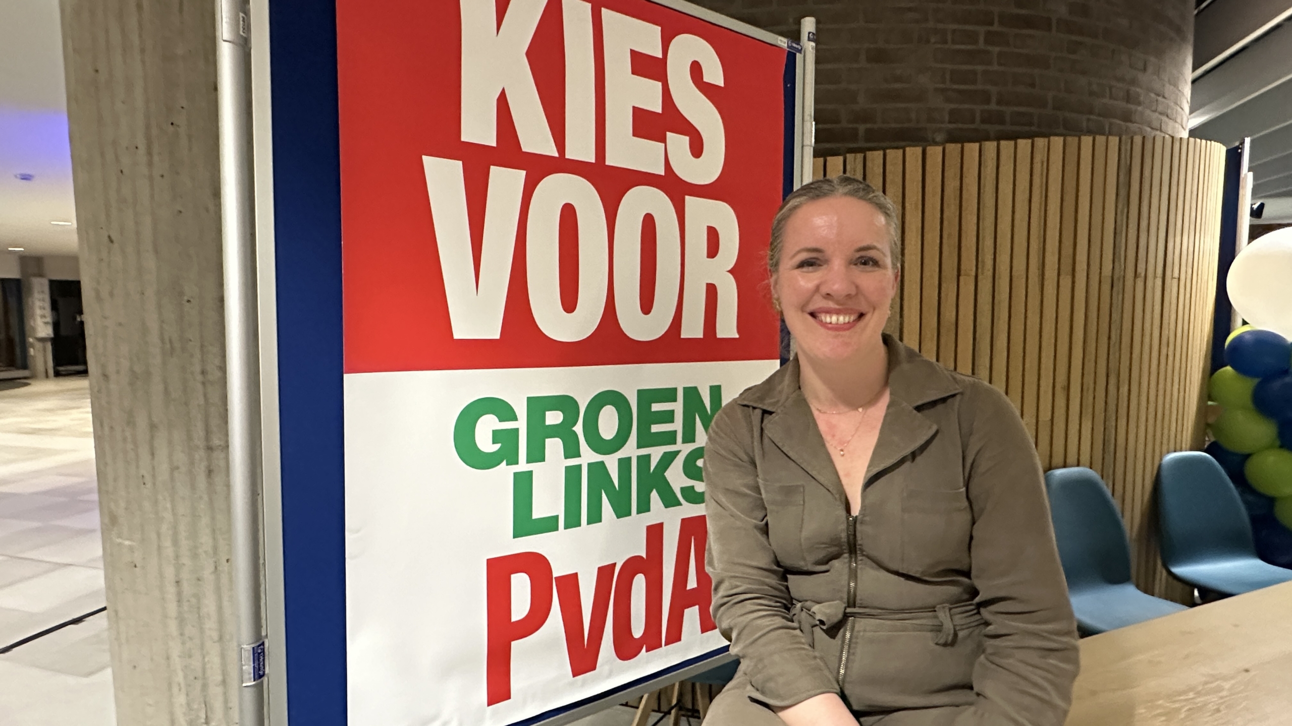 Vrouw glimlacht naast een bord met de tekst "Kies voor GroenLinks PvdA" in een binnenruimte.