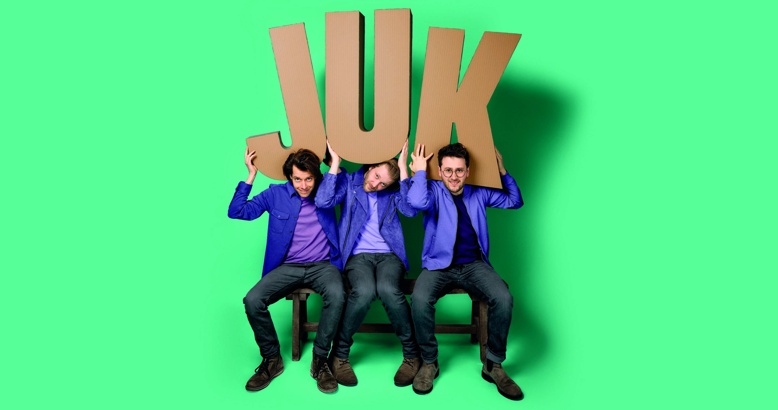 Drie mannen in blauwe jasjes en paarse shirts houden grote letters "JUK" boven hun hoofd tegen een groene achtergrond.