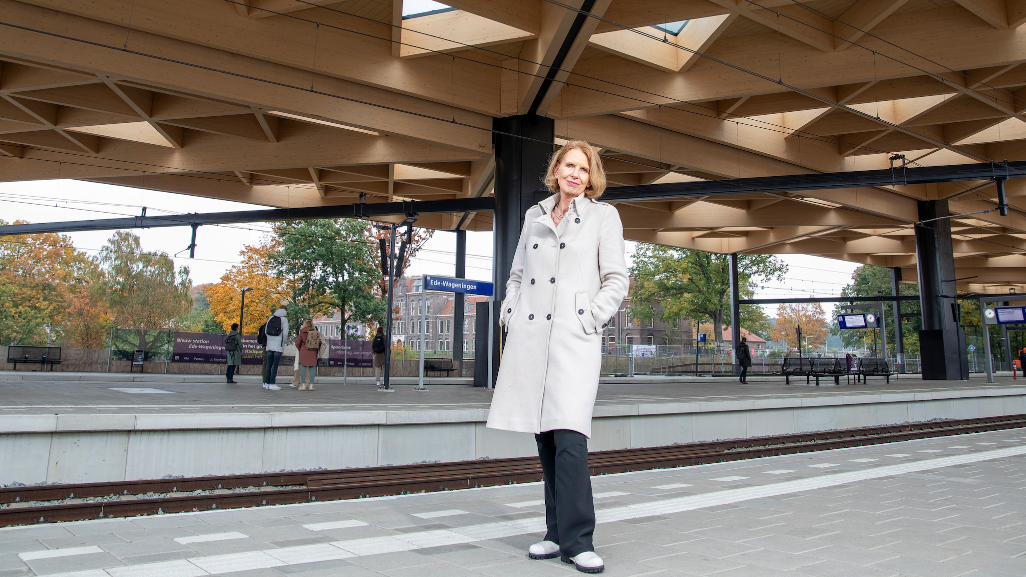 Een vrouw in een witte jas staat op een perron van station Ede-Wageningen, onder een modern houten dak.