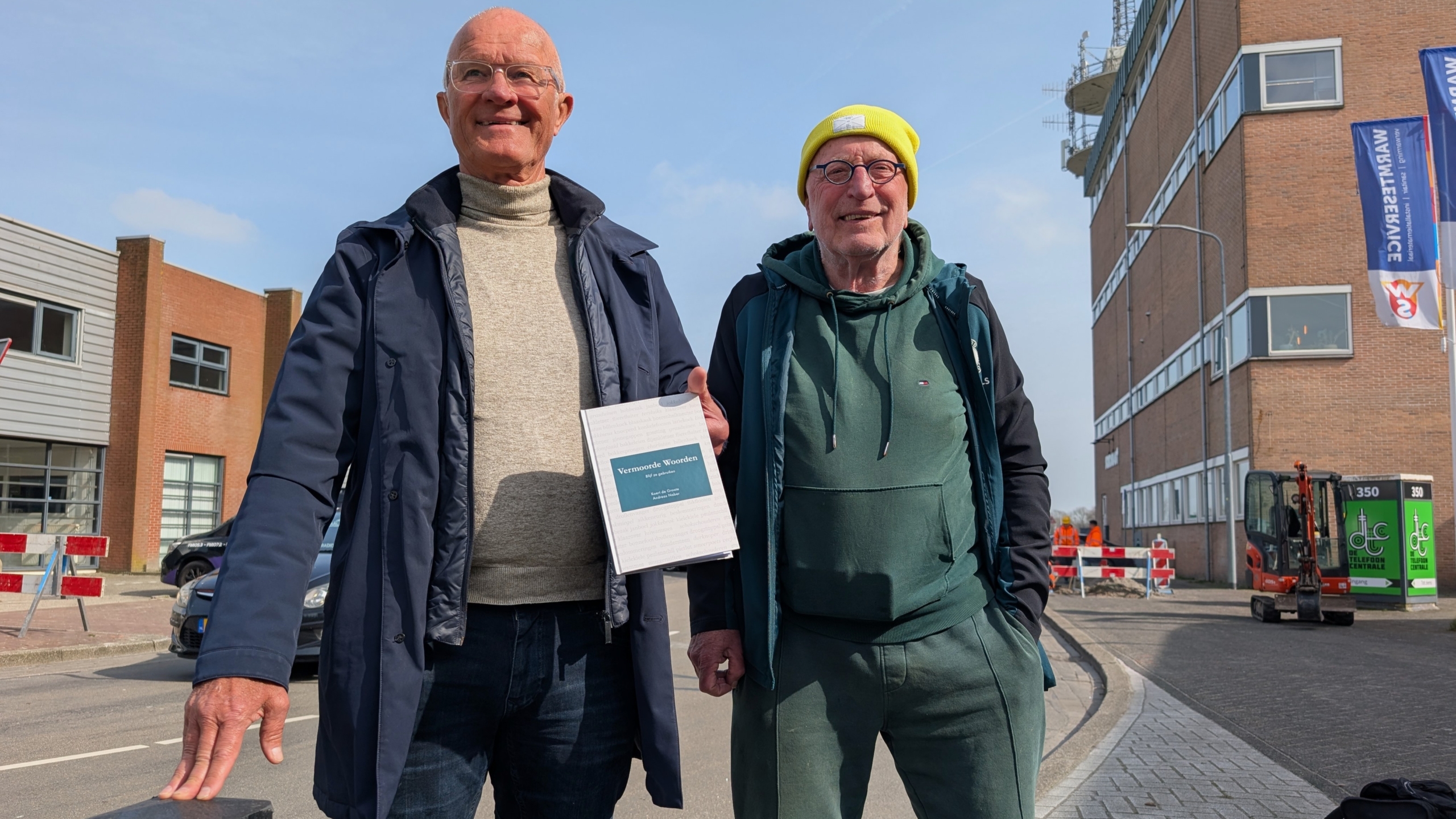 Twee oudere mannen staan op straat, een van hen houdt een boek met de titel "Vermoeide Woorden" vast.