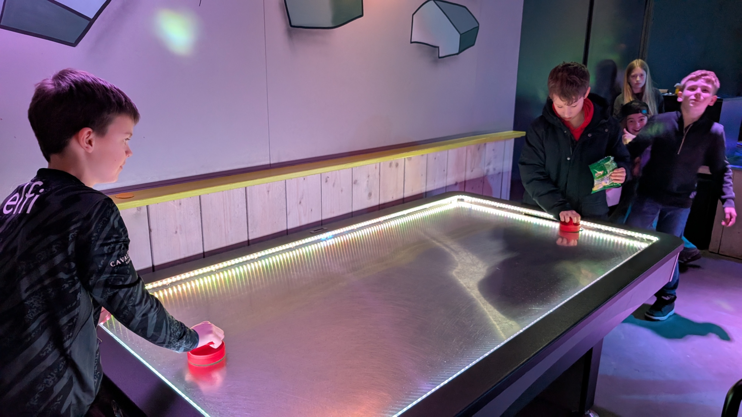 Twee jongens spelen airhockey op een verlicht tafelblad, terwijl anderen toekijken.
