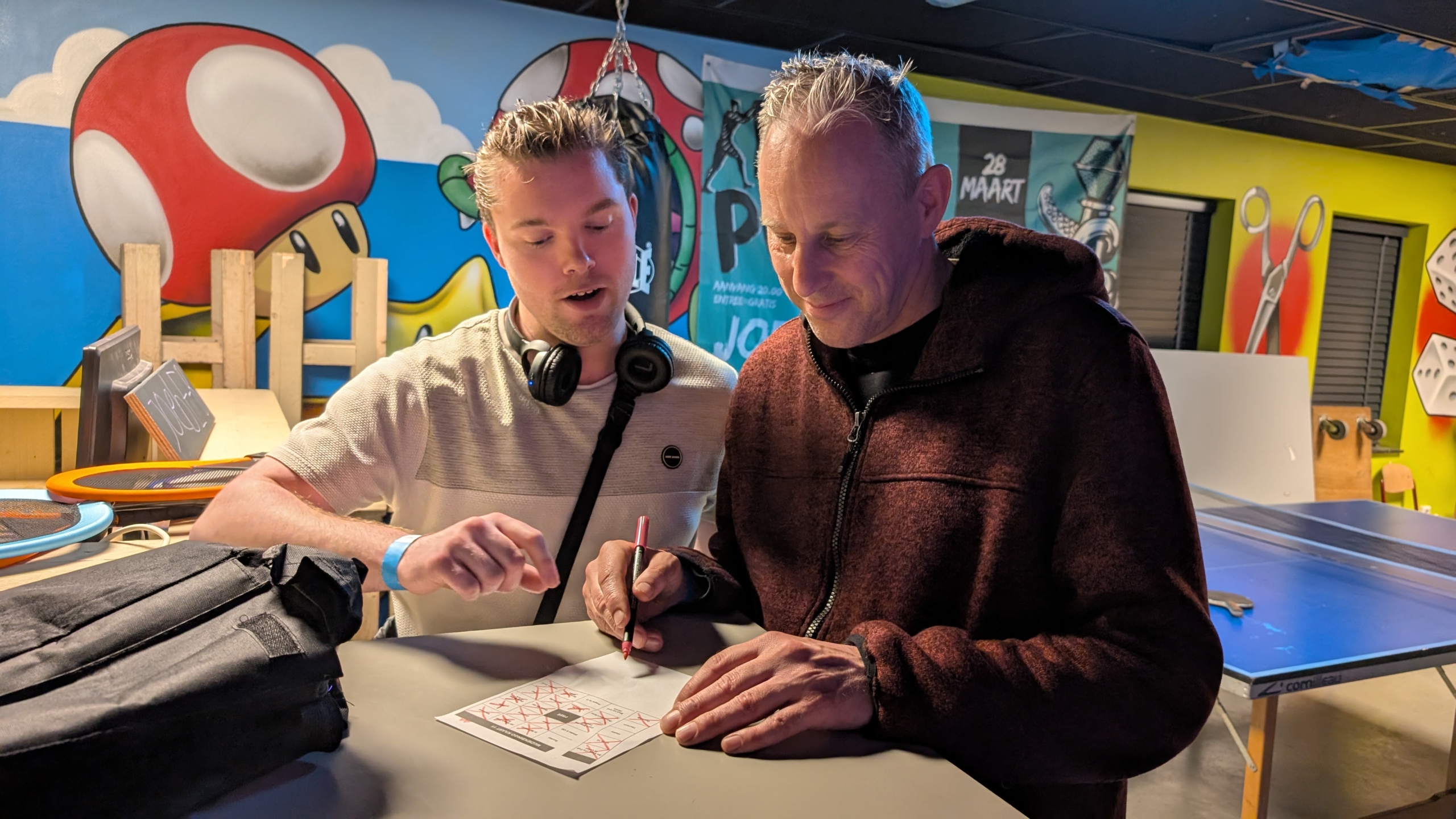 Twee mannen zijn geconcentreerd bezig met een papier op een tafel in een kleurrijke ruimte met muurschilderingen.