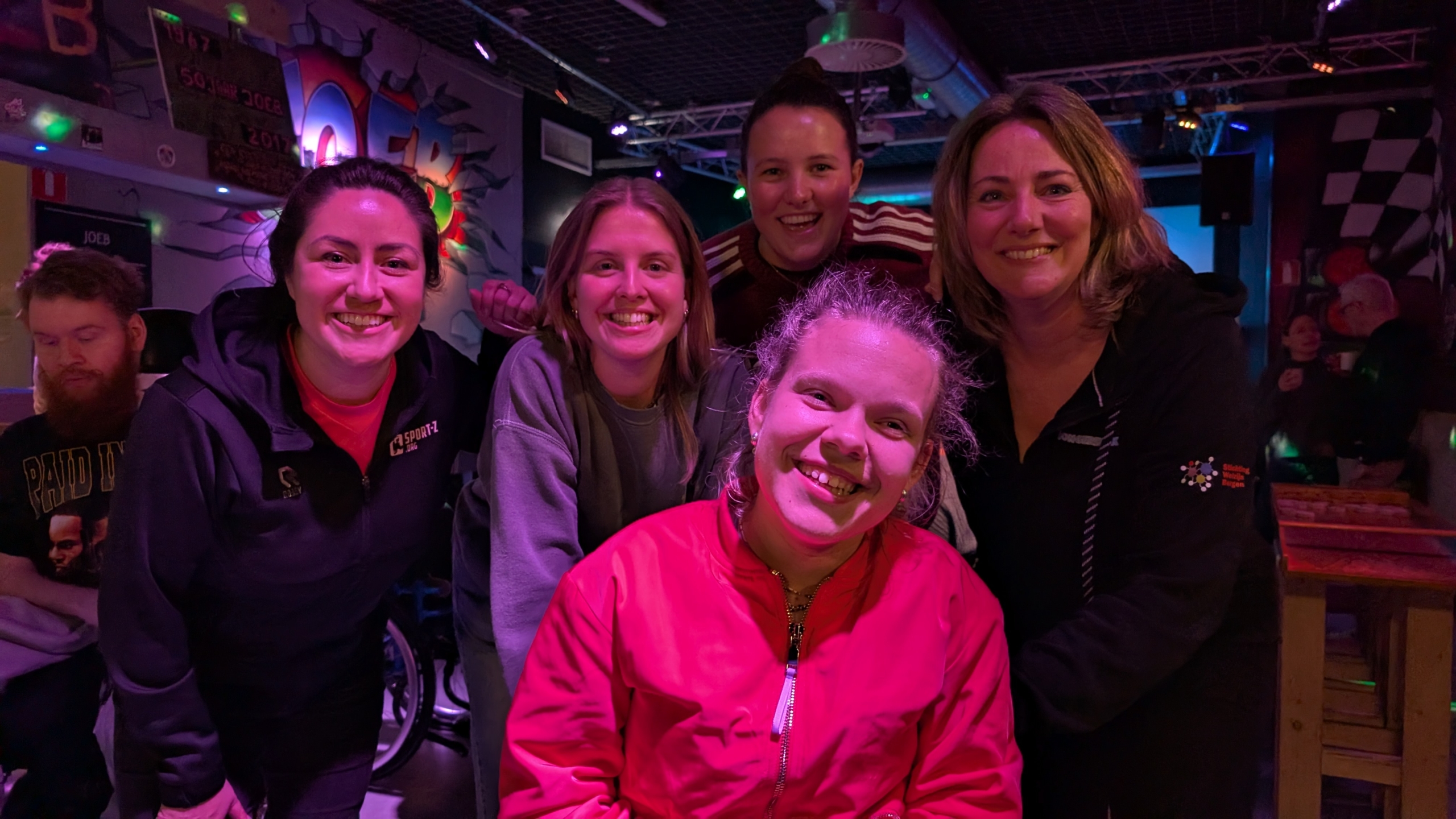 Vijf lachende vrouwen poseren samen in een kleurrijk verlichte ruimte met kunst aan de muur.