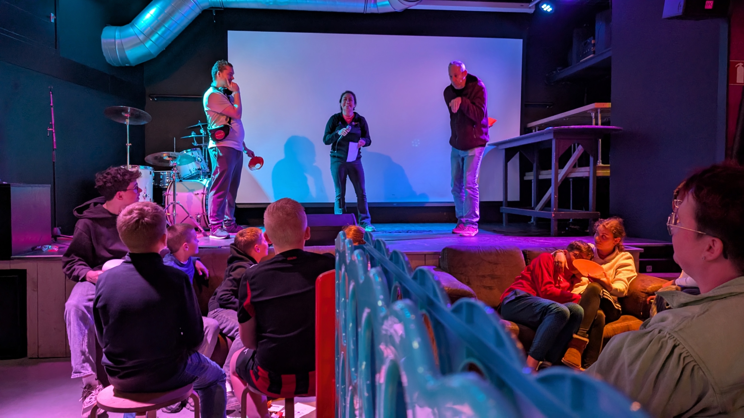 Mensen op een kleine podiumvoorstelling, omringd door een zittend publiek in een kleurrijke ruimte met lichteffecten.