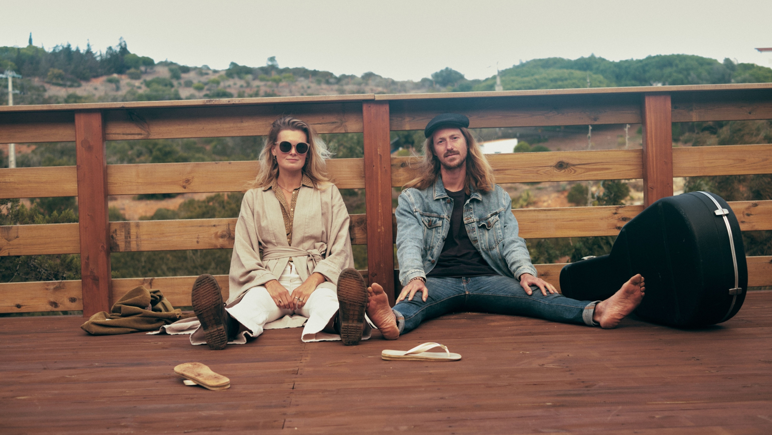 Twee mensen zitten ontspannen op een houten terras, met een gitaarkoffer naast hen en een natuurlijke achtergrond.