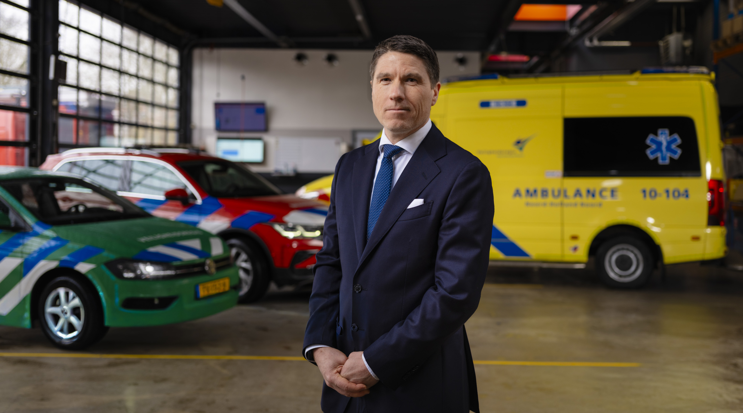 Een man in een donker pak staat in een garage met een groene, een rode auto en een gele ambulance op de achtergrond.