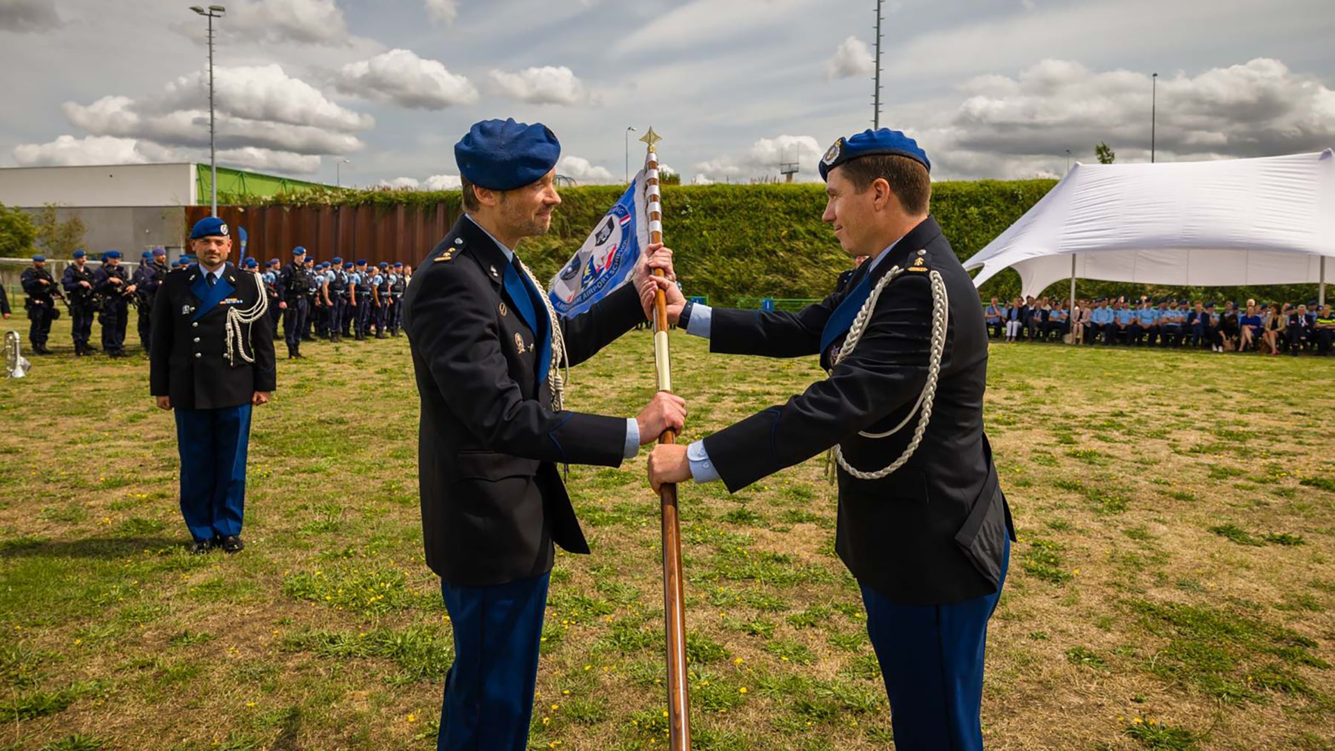 Twee militairen in uniform houden een ceremonieel vaandel vast op een grasveld, met een wachtende groep soldaten en een tent op de achtergrond.