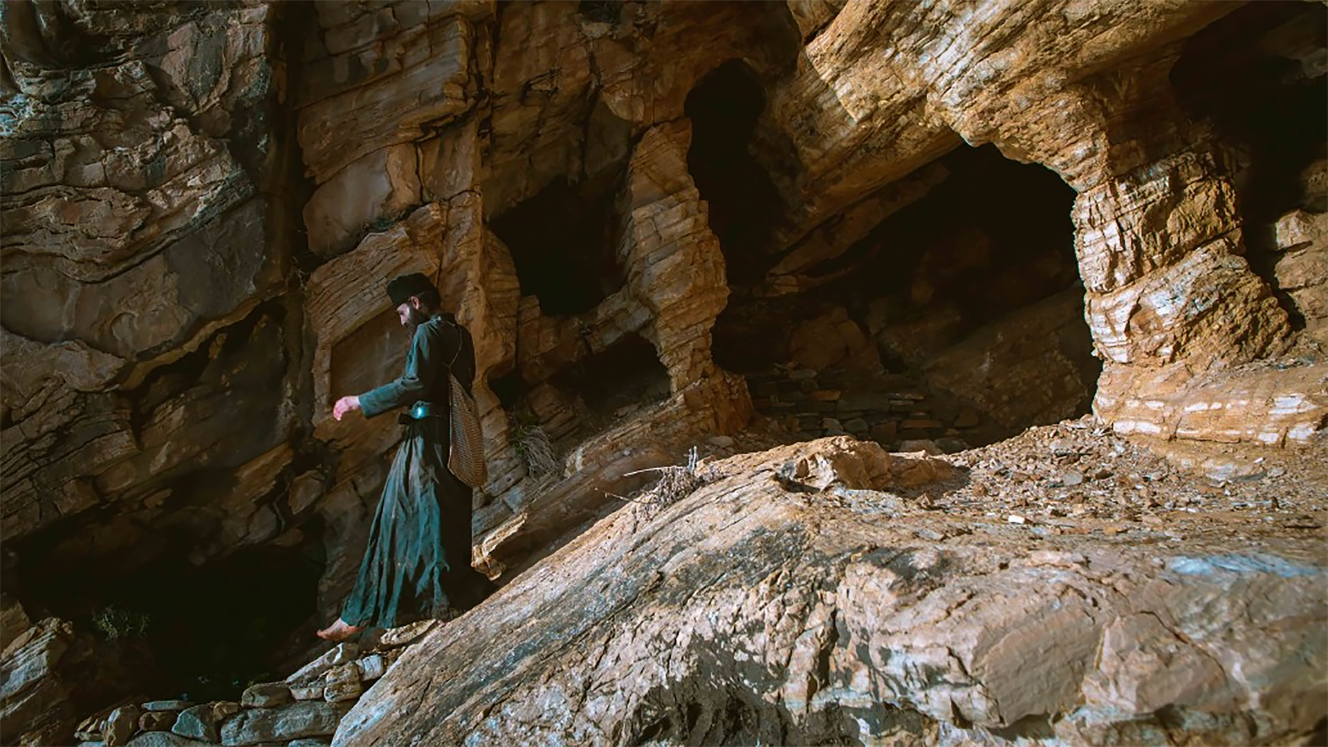 Een persoon loopt langs een rotswand met grotten, gekleed in donkere kleding en een tas dragend.