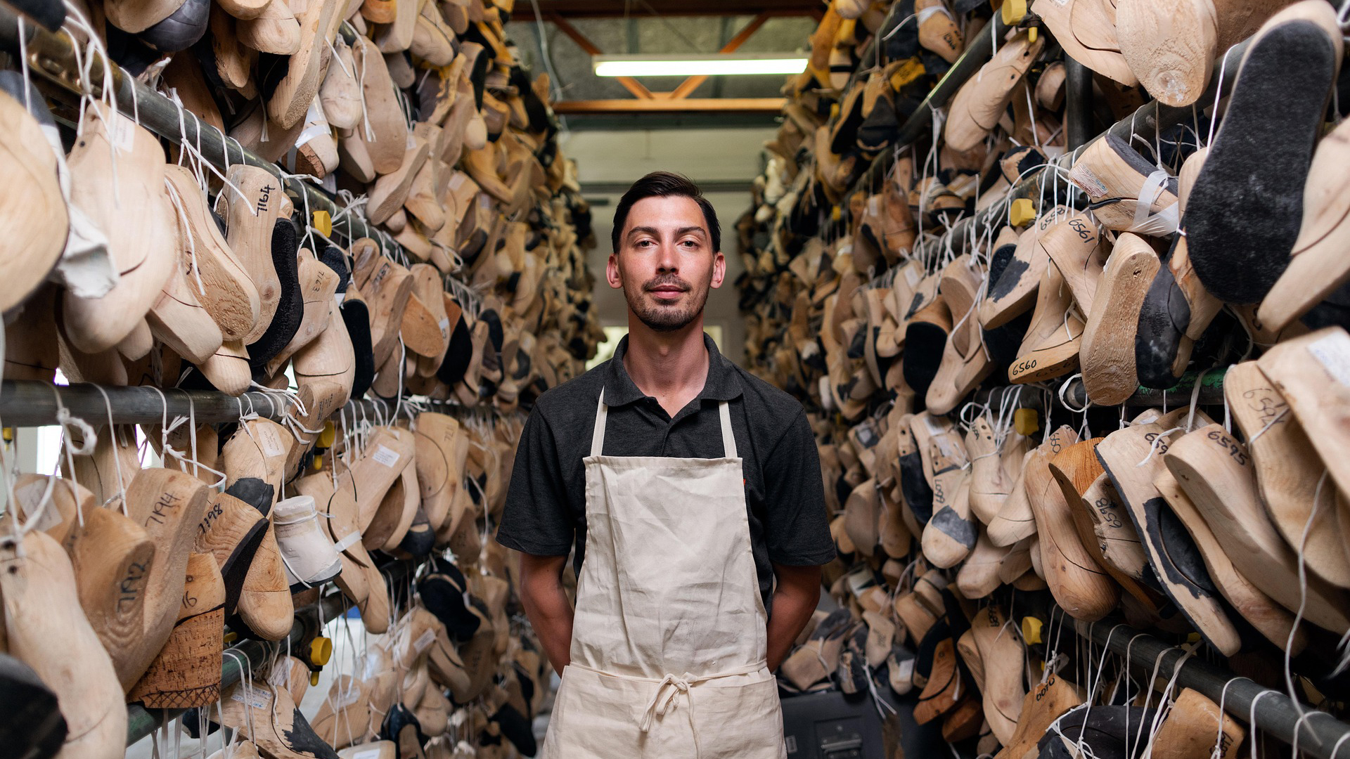 Man met een schort staat tussen rijen houten schoenvormen in een werkplaats.