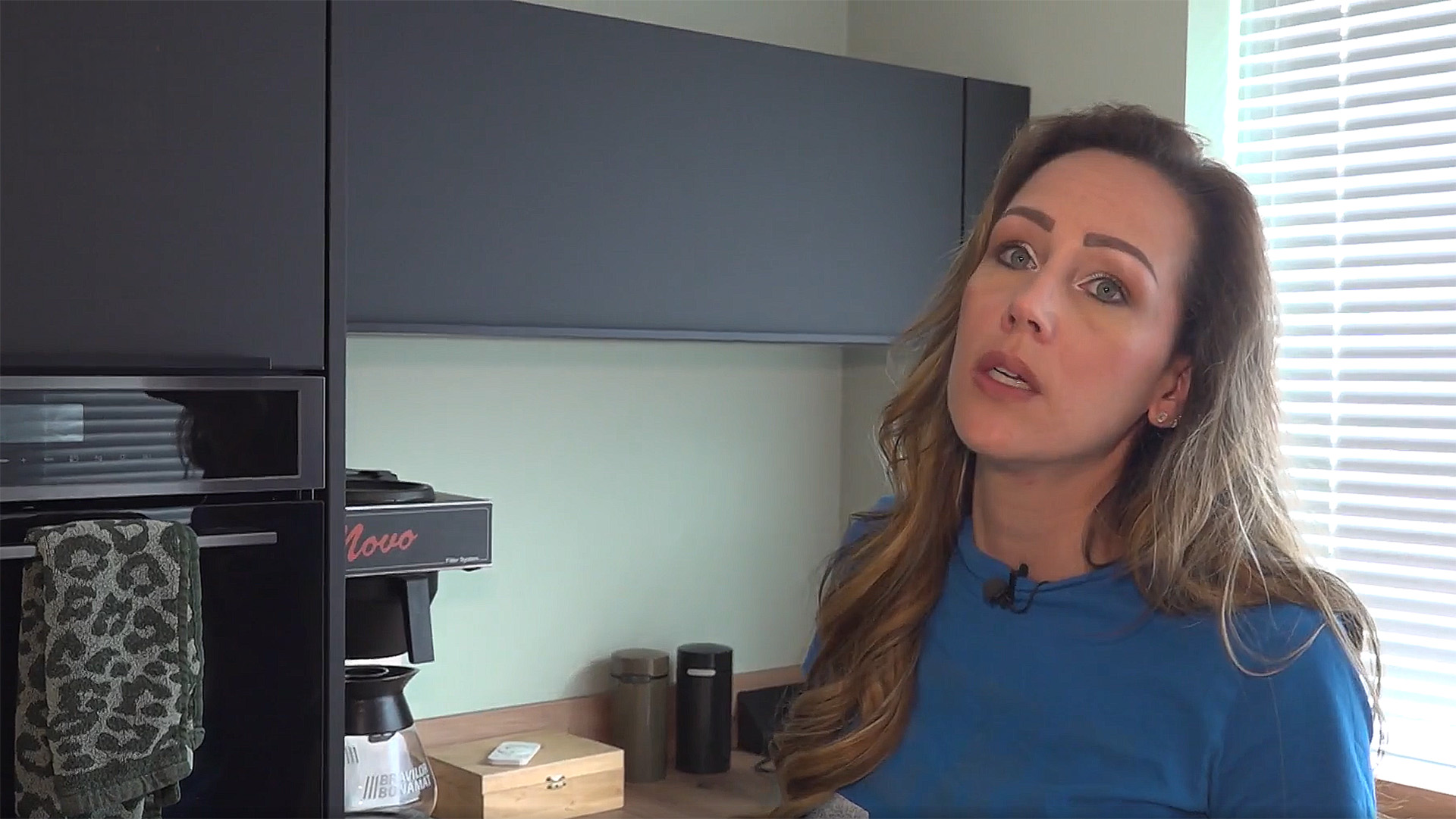 Vrouw in een keuken met een koffiezetapparaat en zwarte kastjes op de achtergrond.