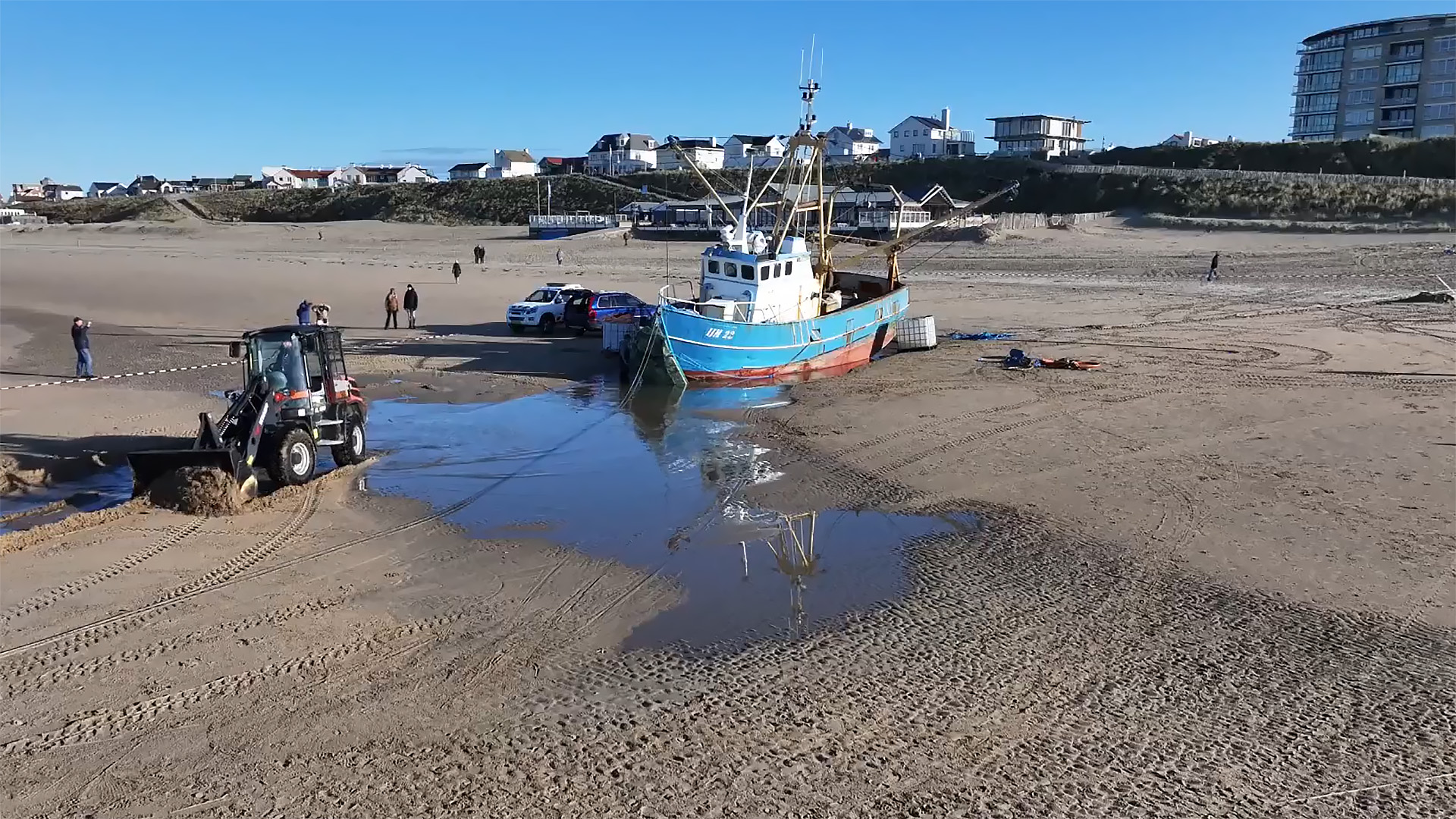Een vissersboot is vastgelopen op een zandstrand, met een tractor en auto's in de buurt.