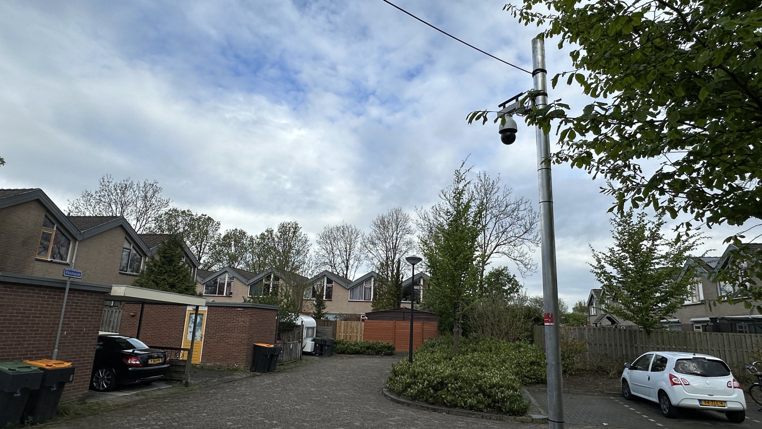 Woonwijk met rijtjeshuizen, geparkeerde auto's, een bewakingscamera op een paal en een halfbewolkte lucht.