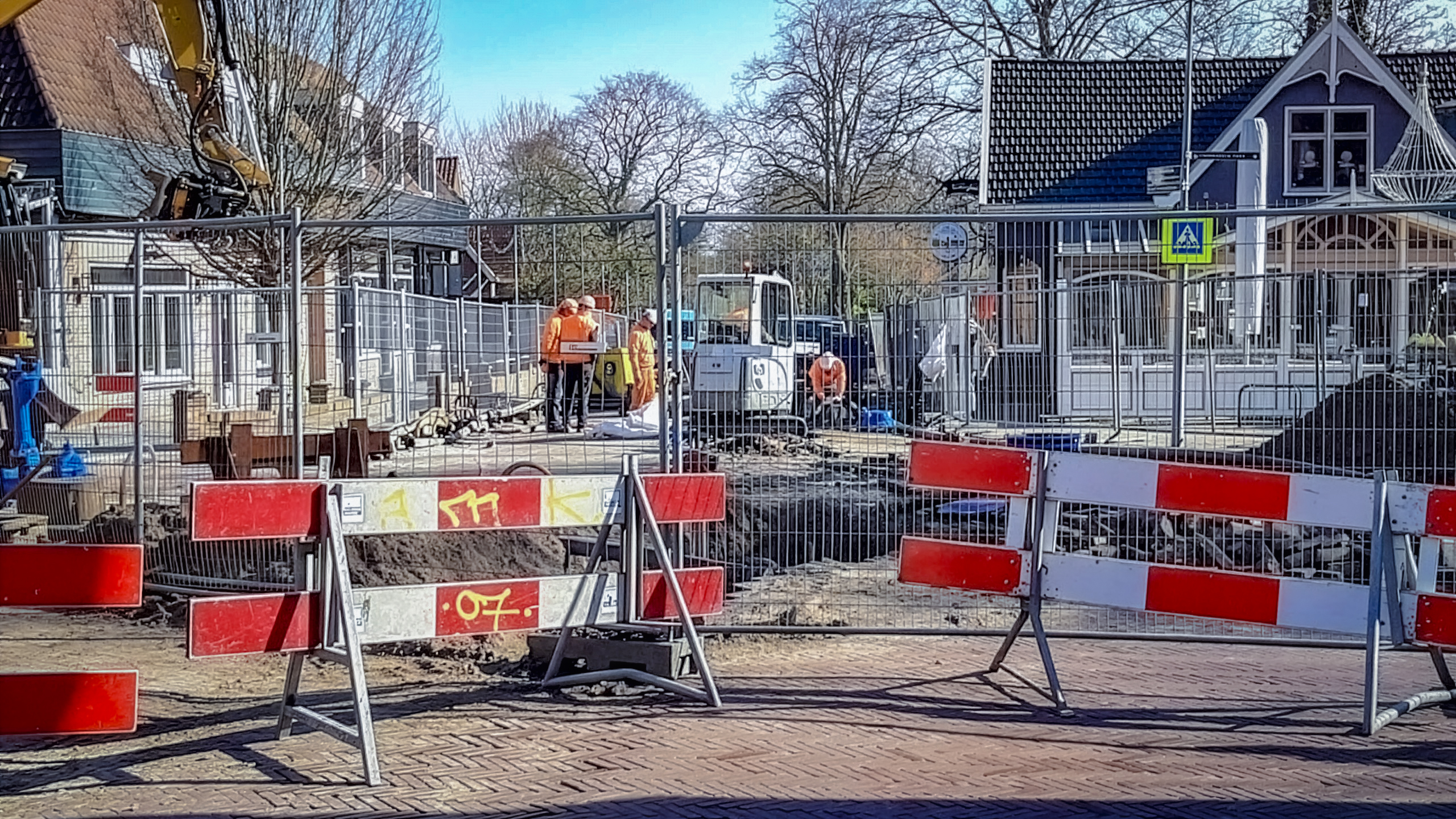 Wegwerkzaamheden met hekken en afzettingen; arbeiders in oranje kleding en een graafmachine op de achtergrond.