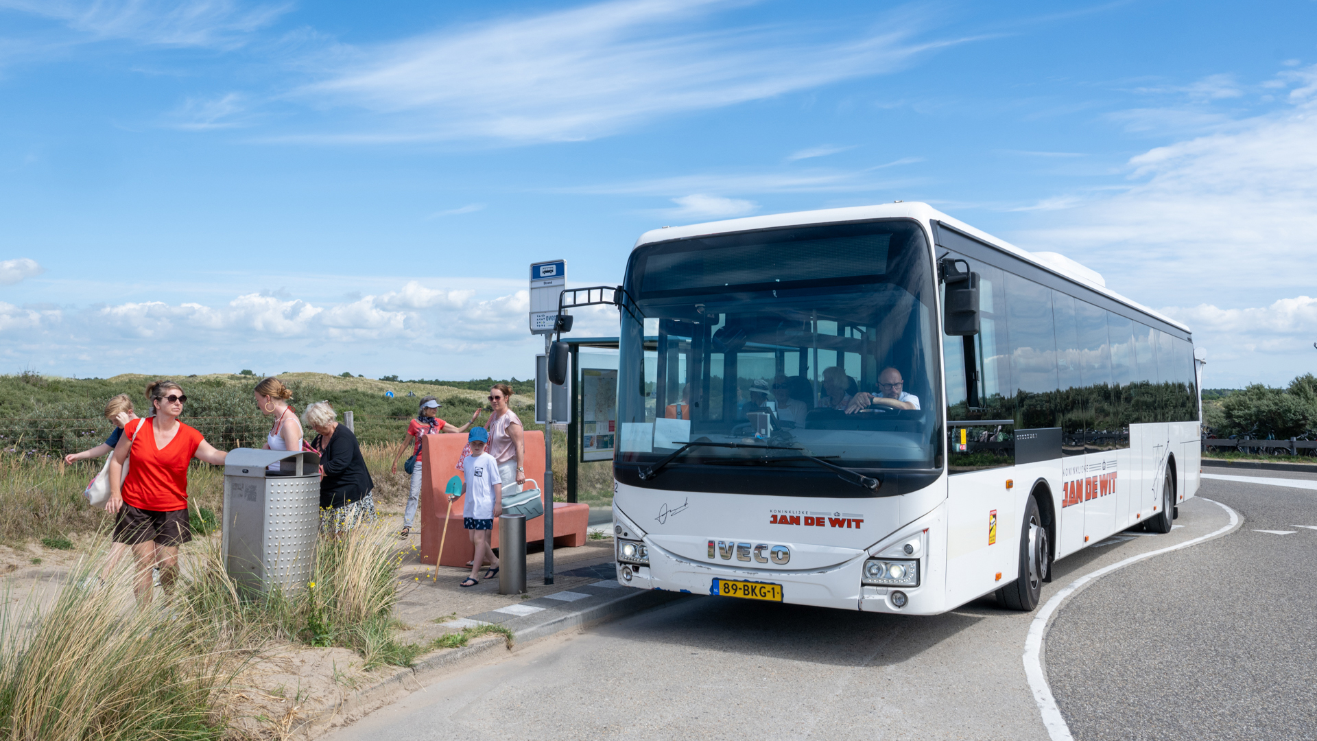 Witte bus met de naam "Jan de Wit" stopt bij een halte in een duinlandschap, omringd door mensen.