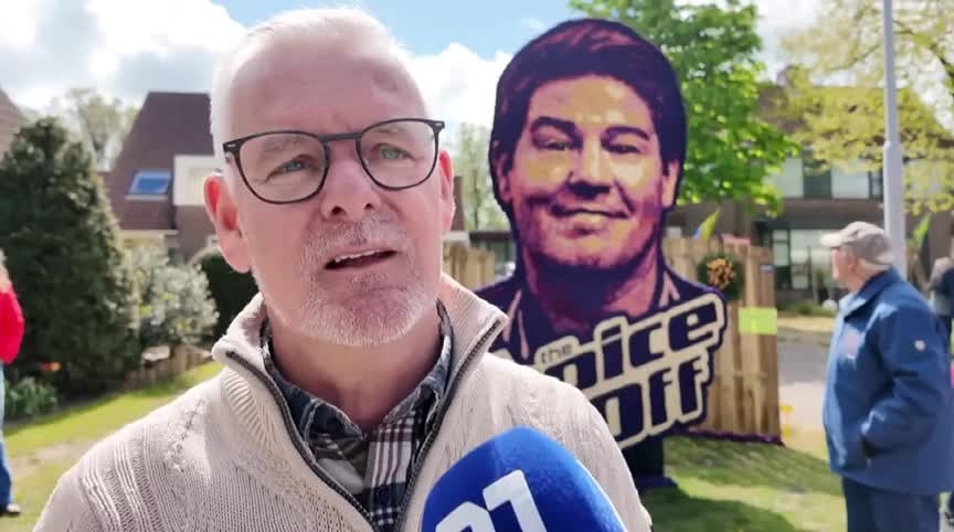 Een man met een bril en een witte baard spreekt in een microfoon buiten, met een groot portret op de achtergrond.