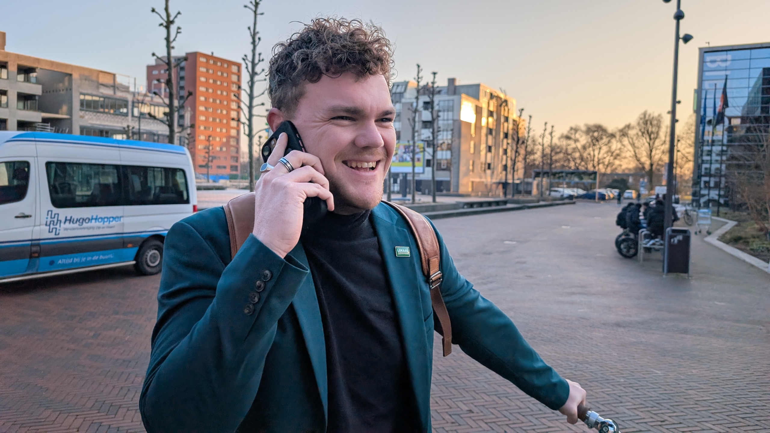 Jonge man met krullend haar lacht en praat aan de telefoon terwijl hij een fiets vasthoudt op een zonnige dag.