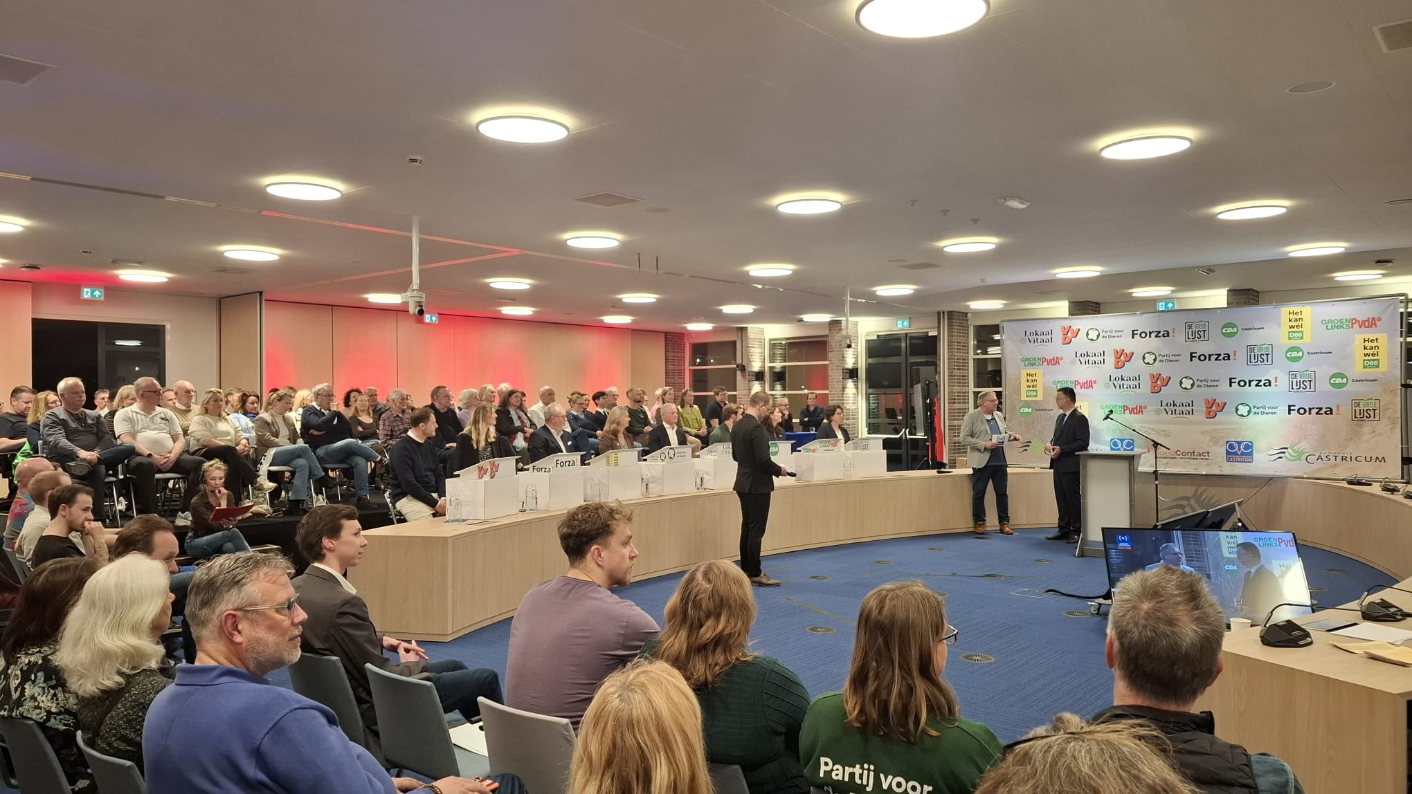 Groep mensen zit in een zaal tijdens een bijeenkomst; twee mannen staan vooraan bij een podium met banners van verschillende politieke partijen.