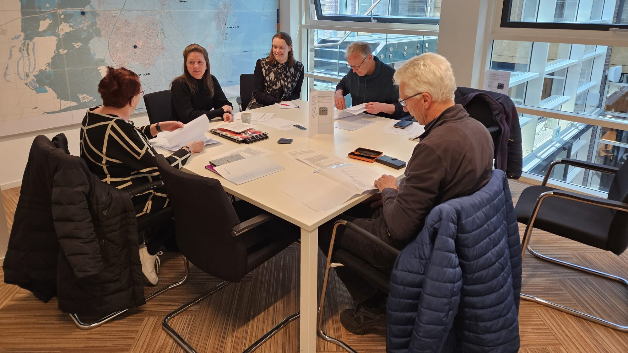 Een groep van zes mensen zit rond een tafel in een vergaderruimte, omgeven door papieren en documenten.