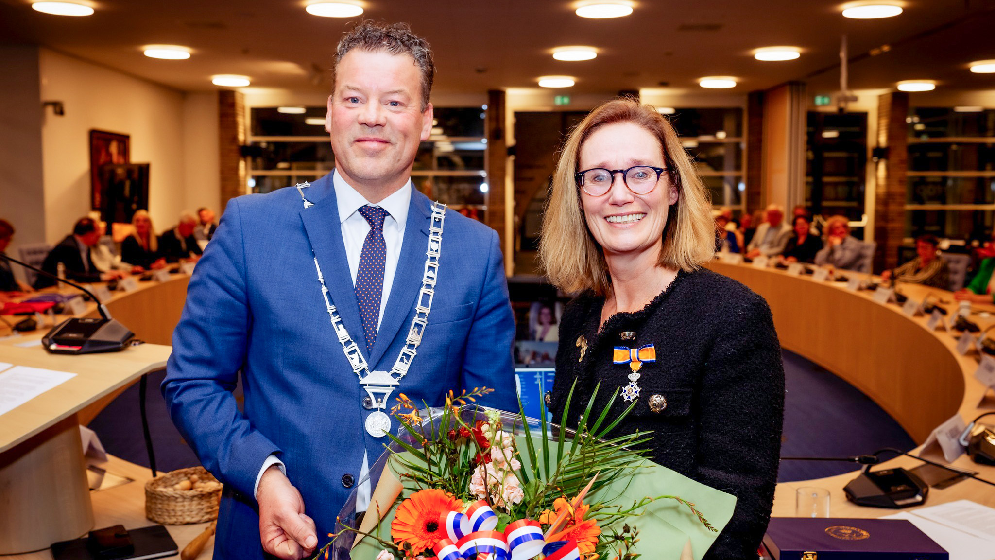 Twee mensen in een formele setting; de vrouw houdt een boeket bloemen vast en draagt een onderscheiding.