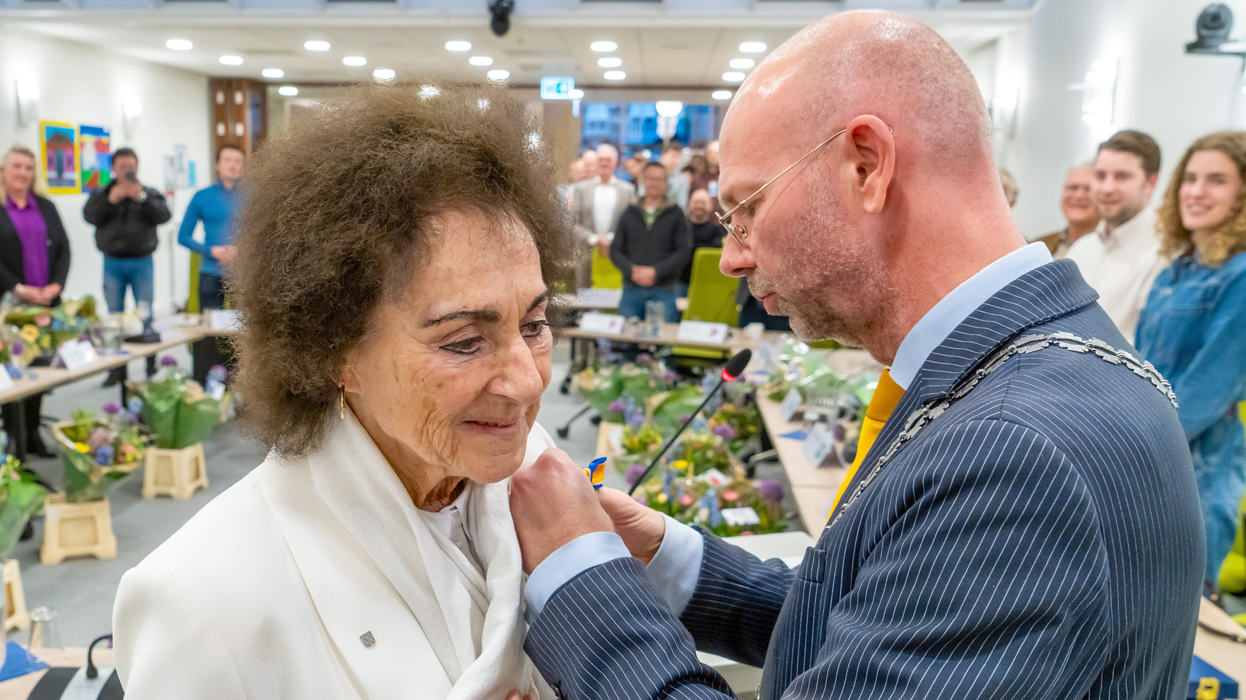 Een man in een pak reikt een onderscheiding uit aan een vrouw in een witte blazer in een zaal met toeschouwers.