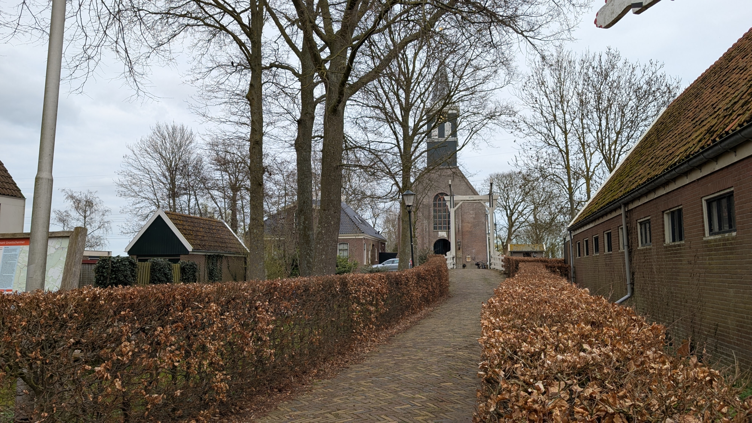 Pad naar een kerkgebouw omringd door kale bomen en gesnoeide heggen, met een oude houten poort op de voorgrond.