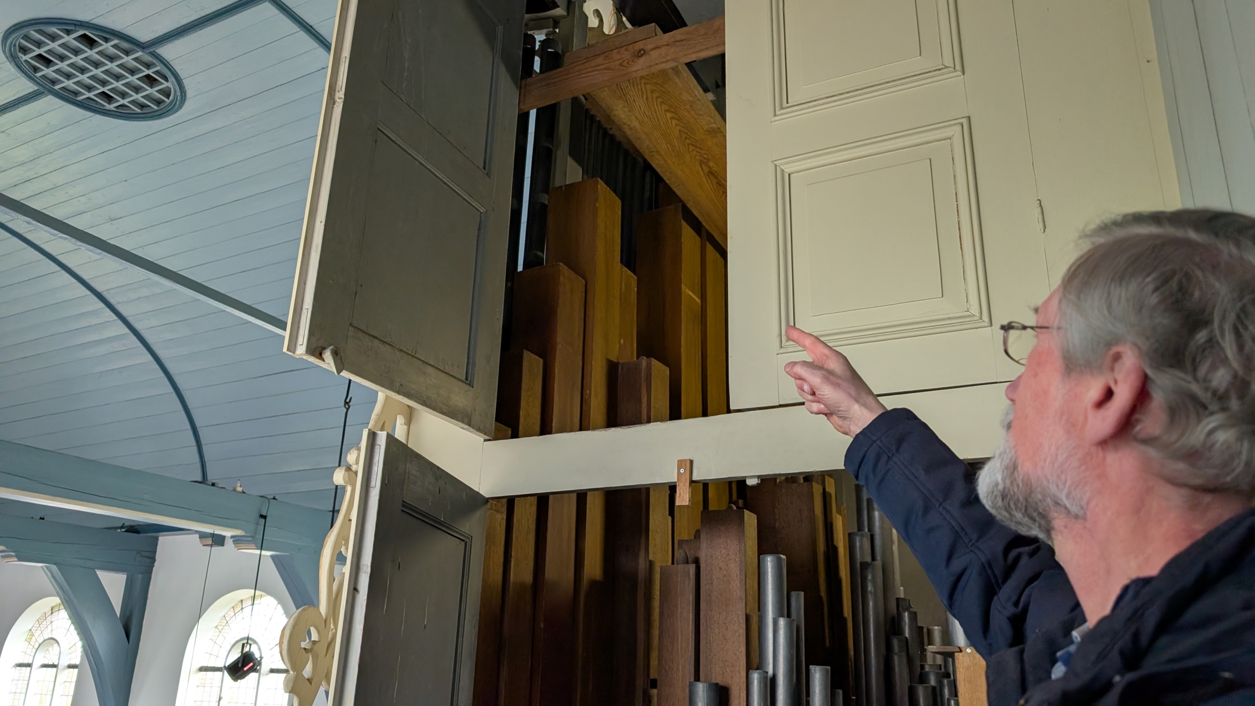 Een man met een bril wijst naar de geopende deuren van een orgelkast met houten orgelpijpen zichtbaar binnenin.