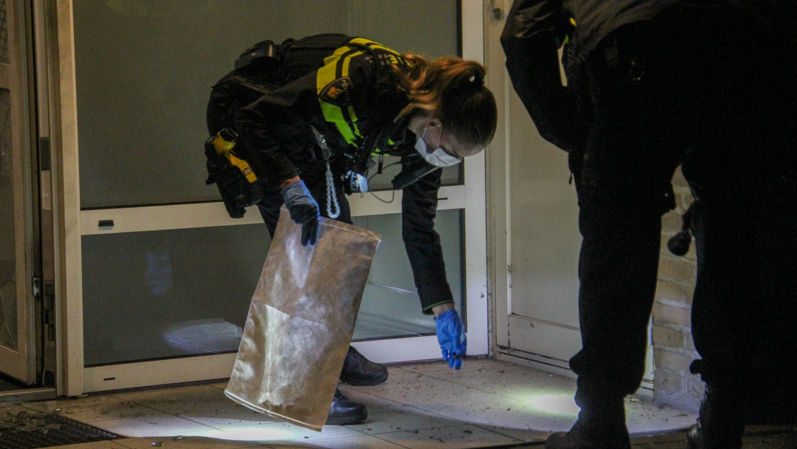Politieagenten onderzoeken een plaats delict bij de ingang van een gebouw, gebruikmakend van een papieren zak en zaklampen.